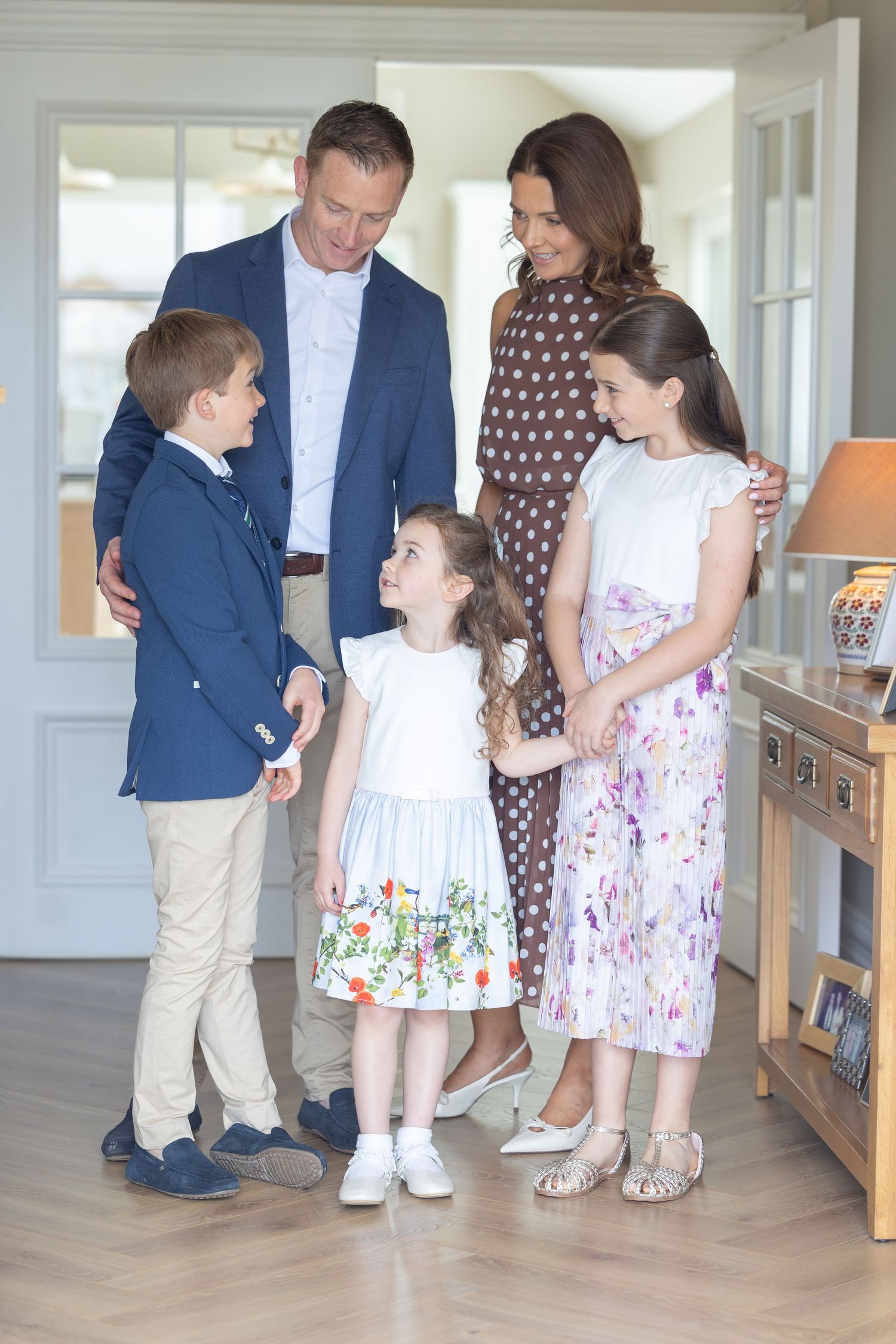 A happy family of five poses indoors, with parents and three children dressed in smart casual spring outfits.