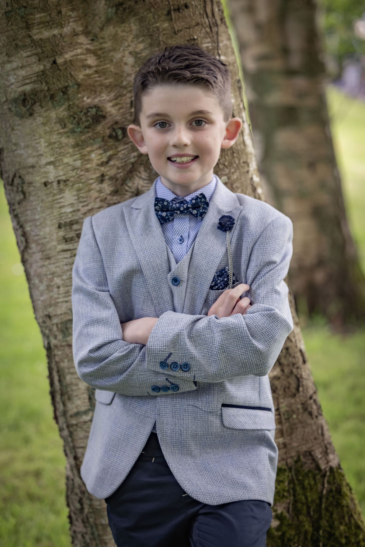 Child in grey suit jacket and polka dot bow tie poses confidently by tree.