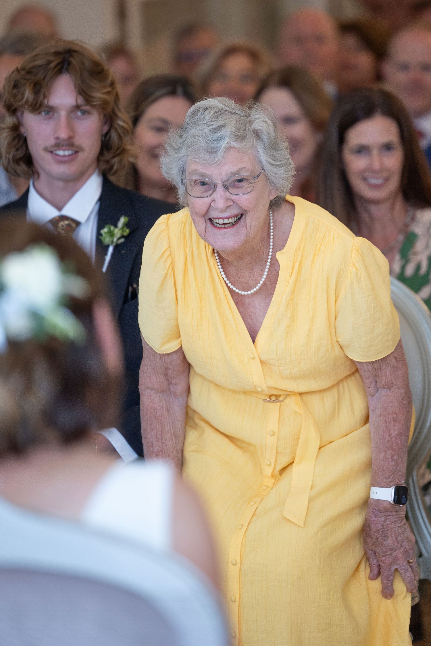 Elderly woman in yellow dress and pearl necklace smiling joyfully at a wedding ceremony surrounded by guests.