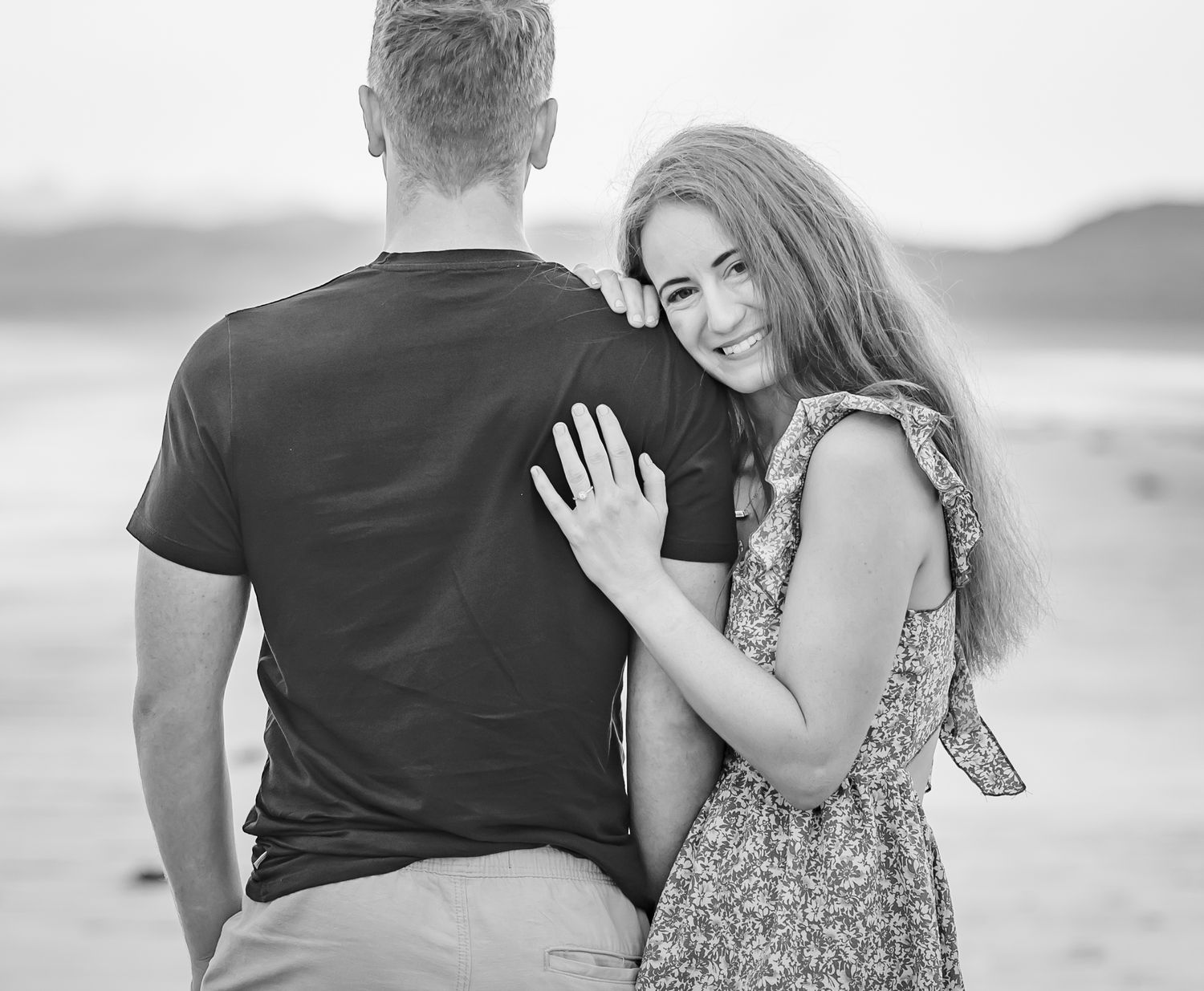 Black and white photo of a couple embracing on a beach, woman smiling over mans shoulder in floral dress.