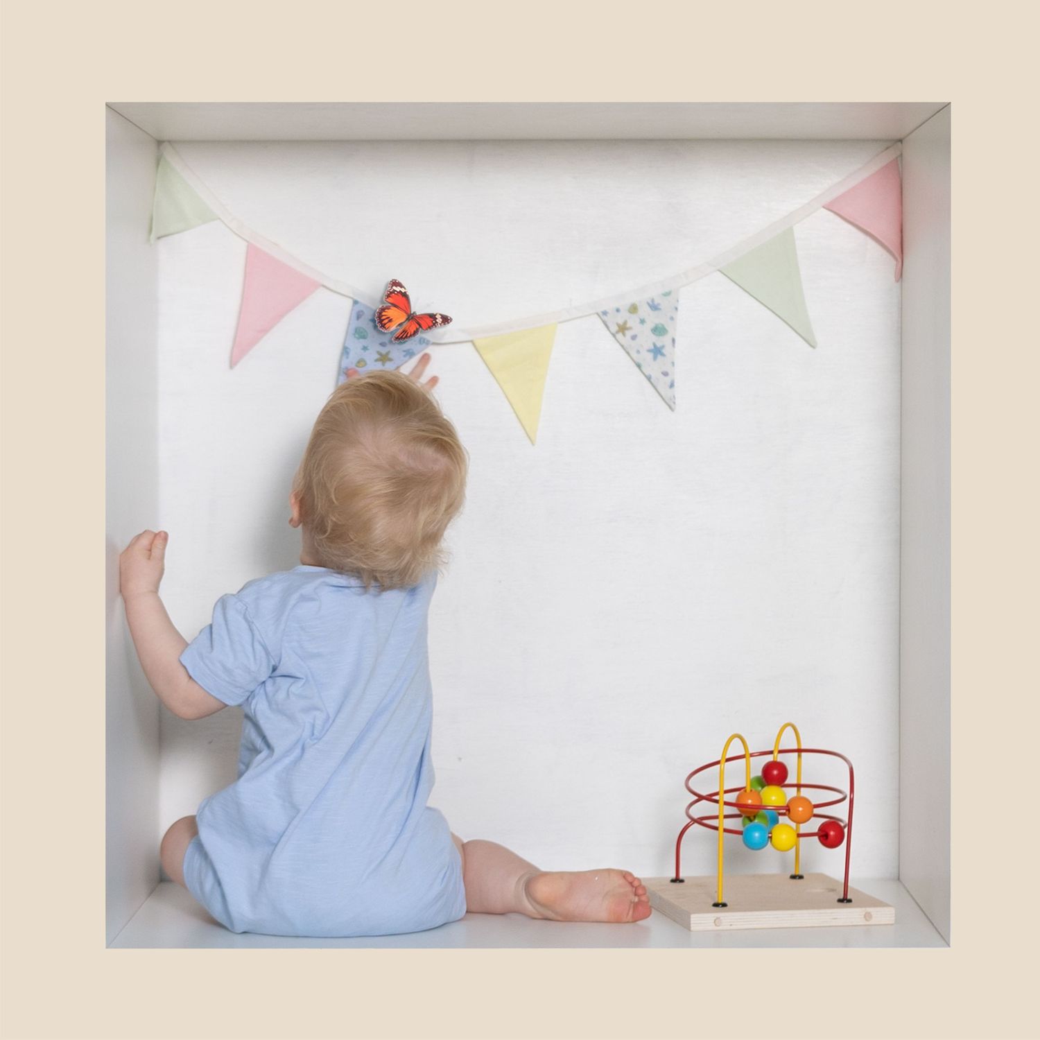 Baby in blue onesie sits facing colorful bunting banner with butterfly, beside a wooden bead maze toy.