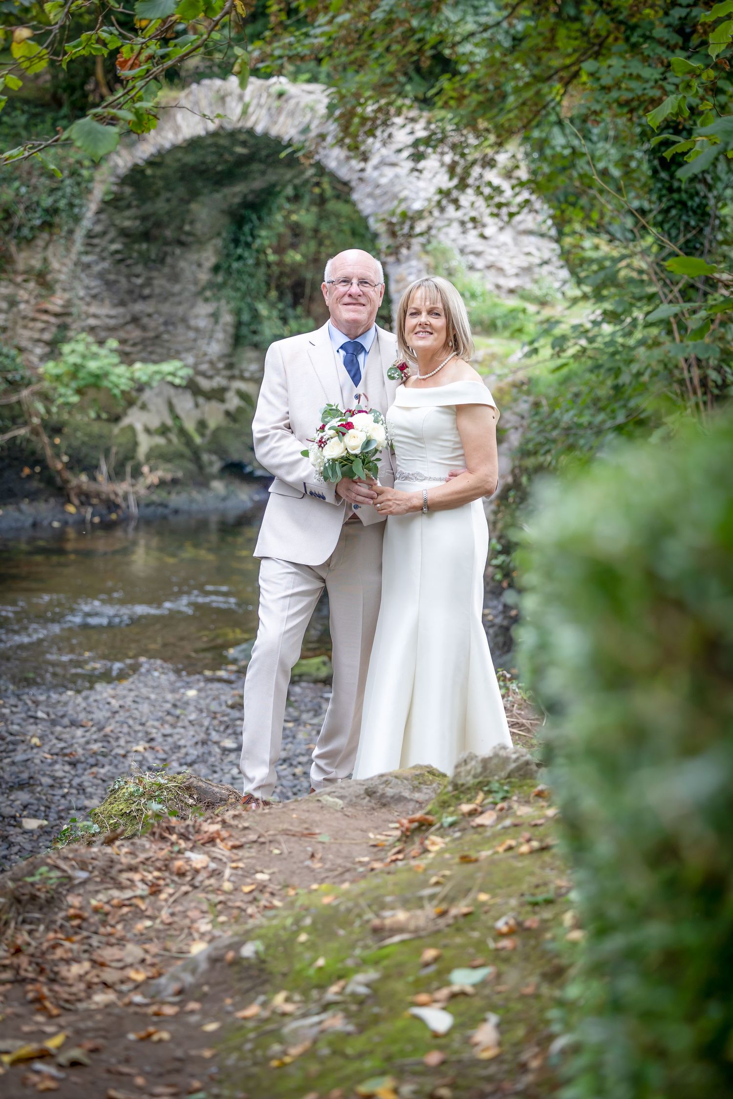 Couple poses in formal attire by stone arch bridge surrounded by greenery.