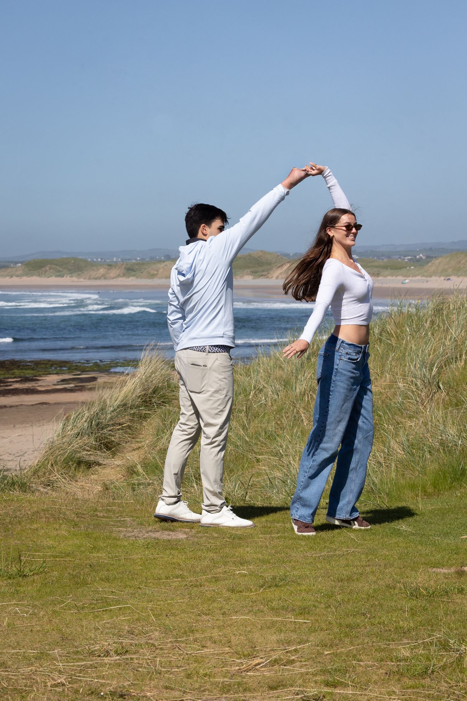 Couple dancing on coastal grass dunes with sandy beach and ocean waves in the background on a sunny day.