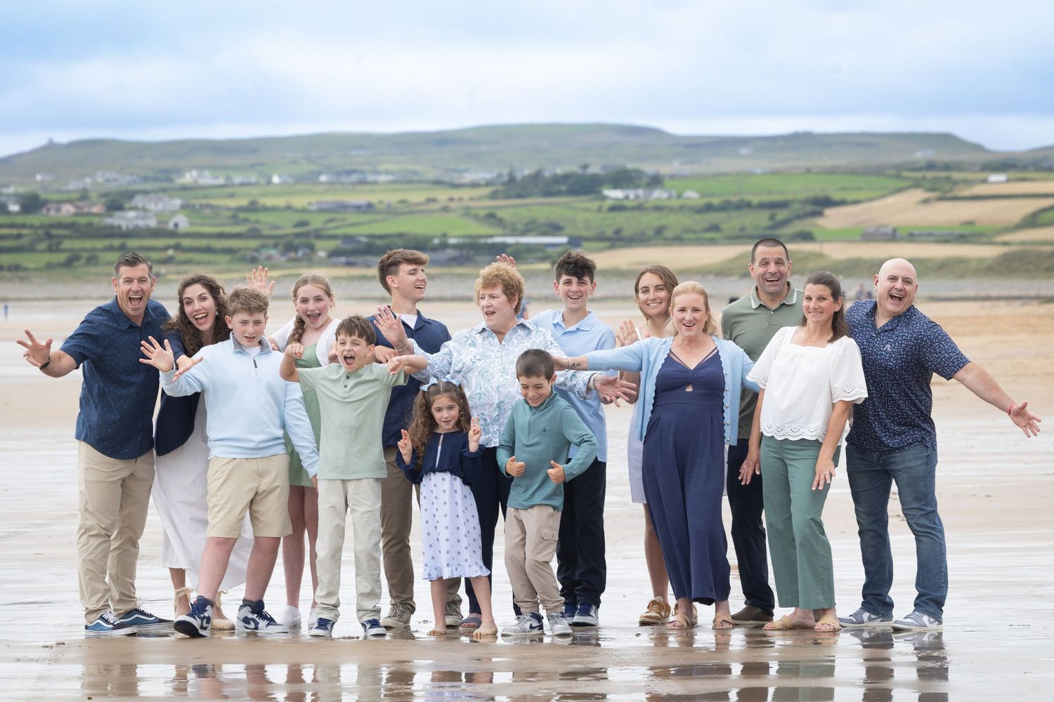 Large family group posing together on a sandy beach with green hills in the background on a cloudy day.