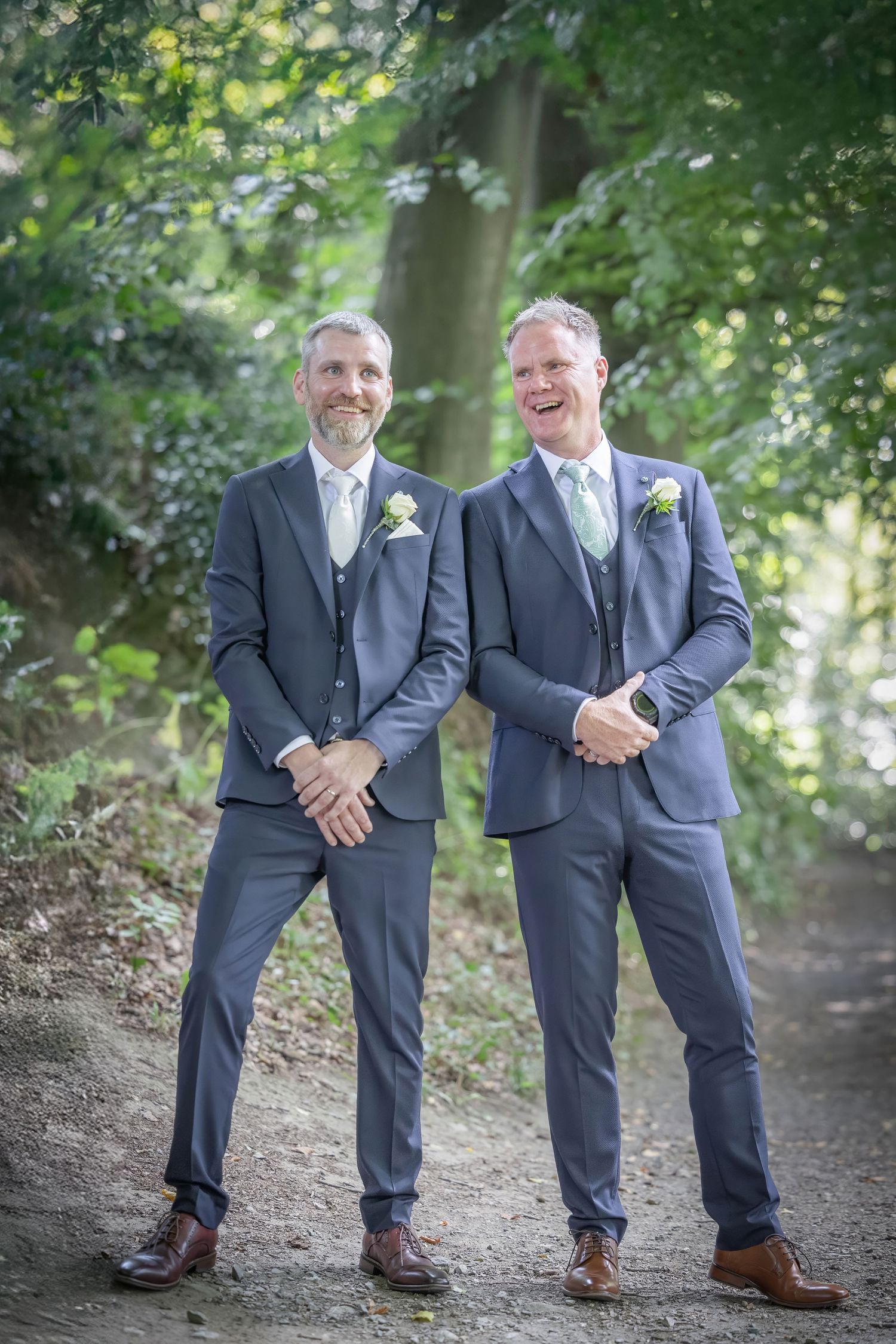 Two men in matching navy suits and ties pose on a forest path.