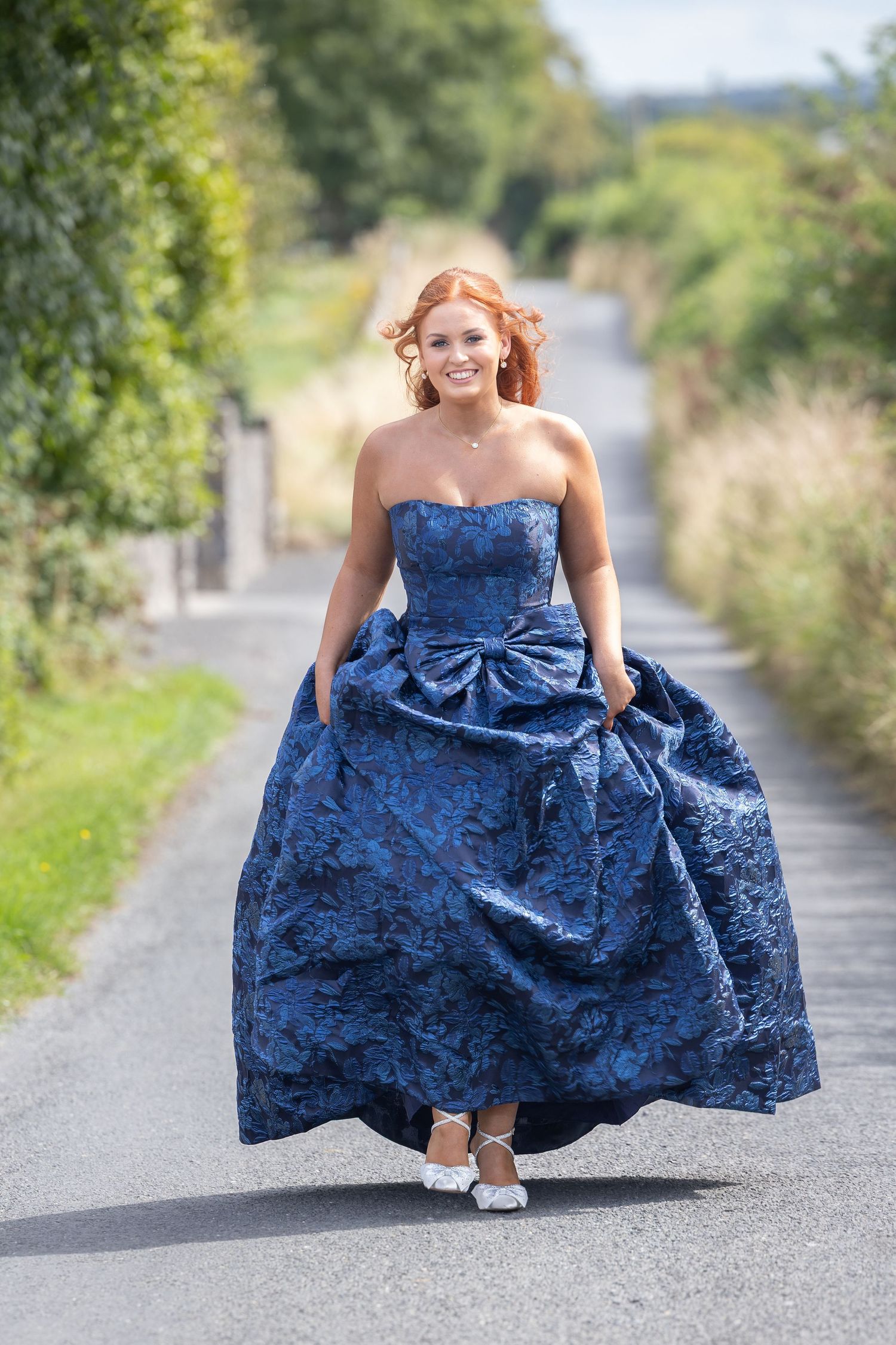 Young woman in elegant blue strapless ball gown walking down a tree-lined country road, smiling.