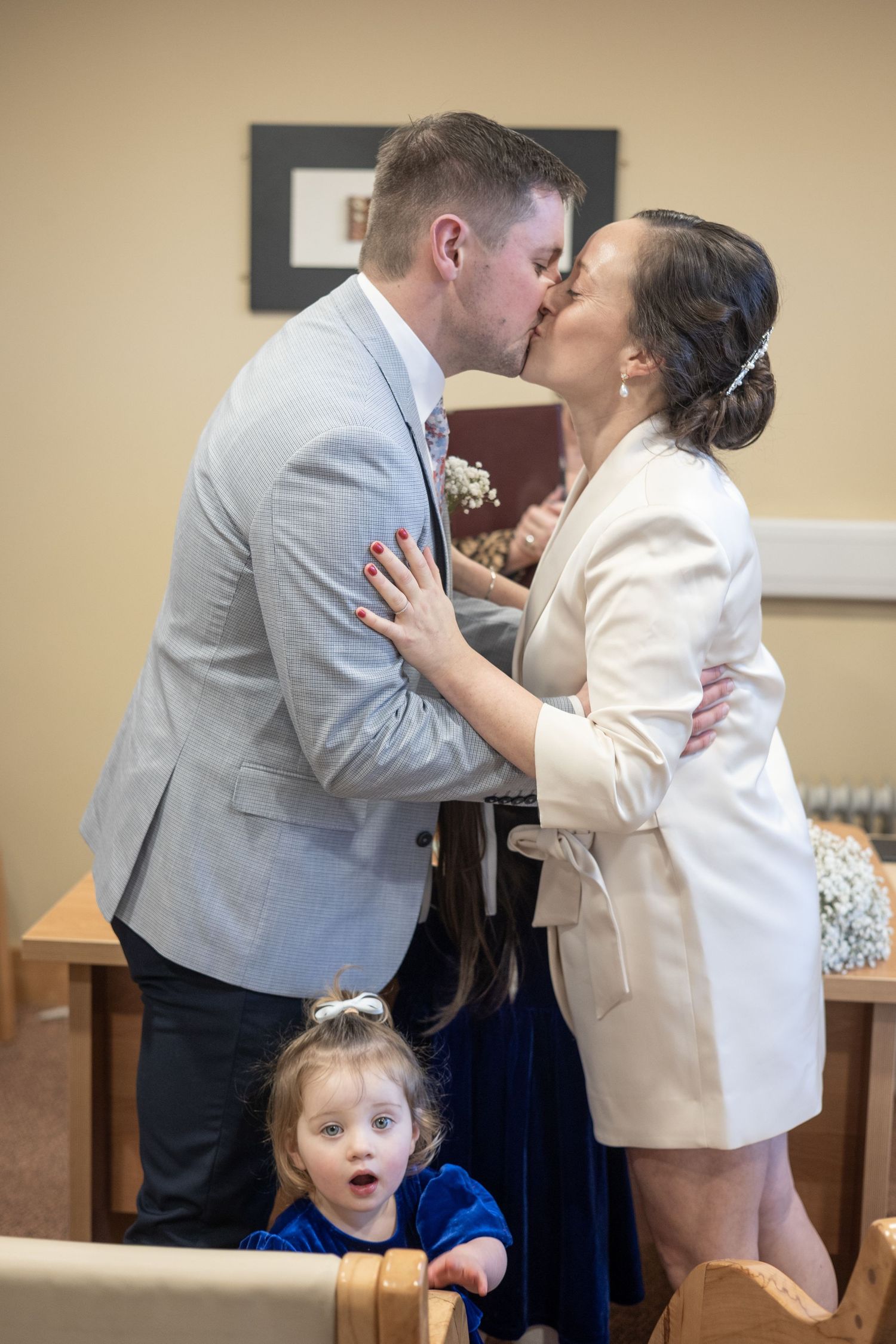 A couple shares a romantic kiss at what appears to be a courthouse while a small child makes a silly face in the foreground.
