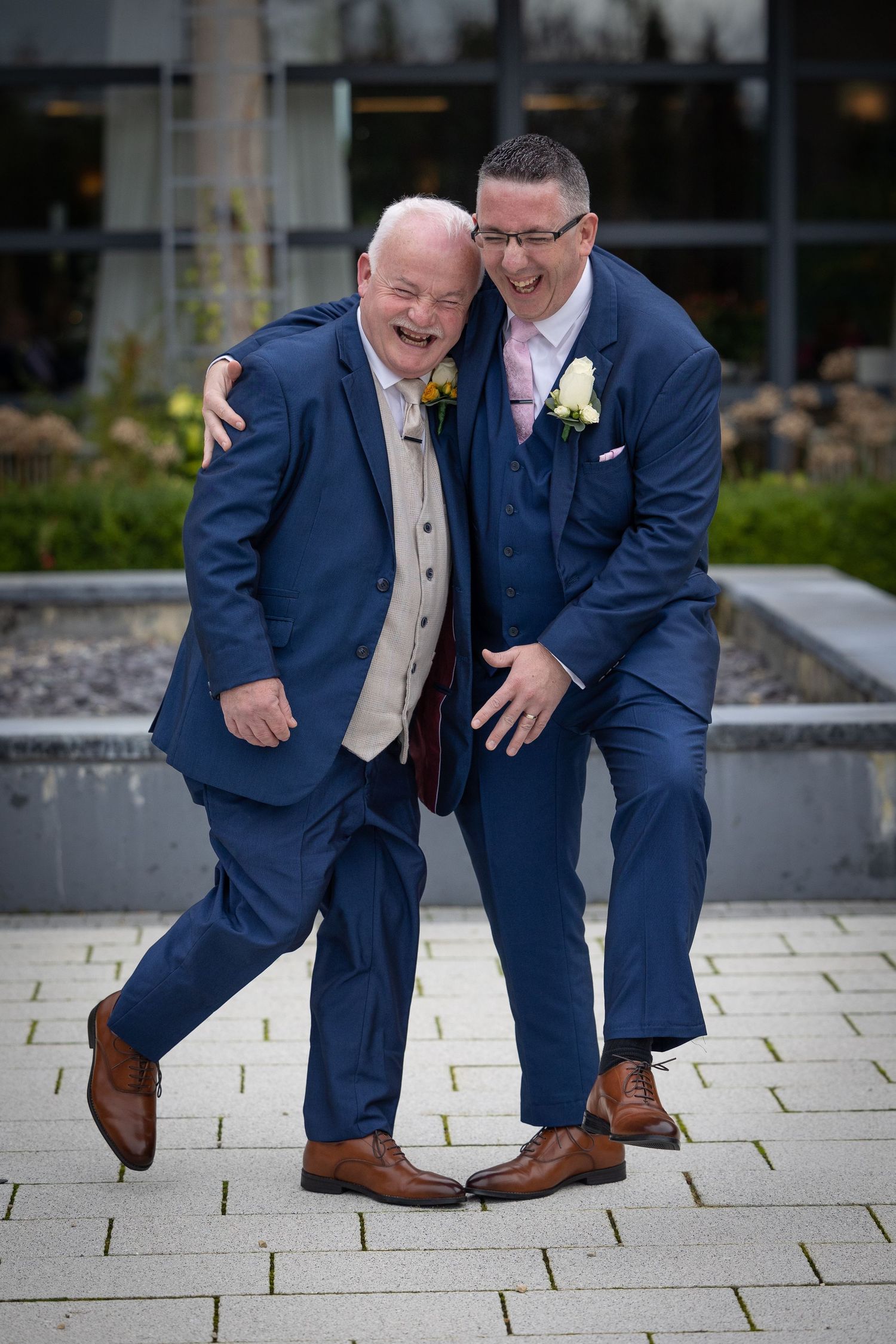 Two people in navy suits and boutonnières share a joyful candid moment together on a stone paved walkway.