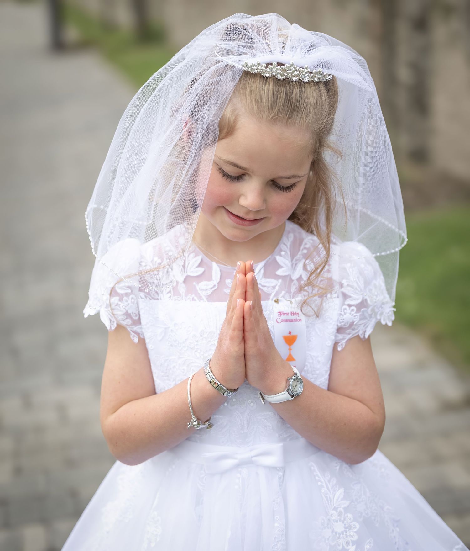 Young girl in white First Communion dress and veil, hands clasped in prayer, smiling downward outdoors.