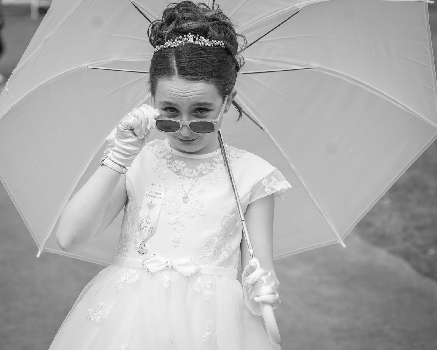 Young girl in white dress and tiara holds umbrella, wearing sunglasses and gloves in black and white photo.
