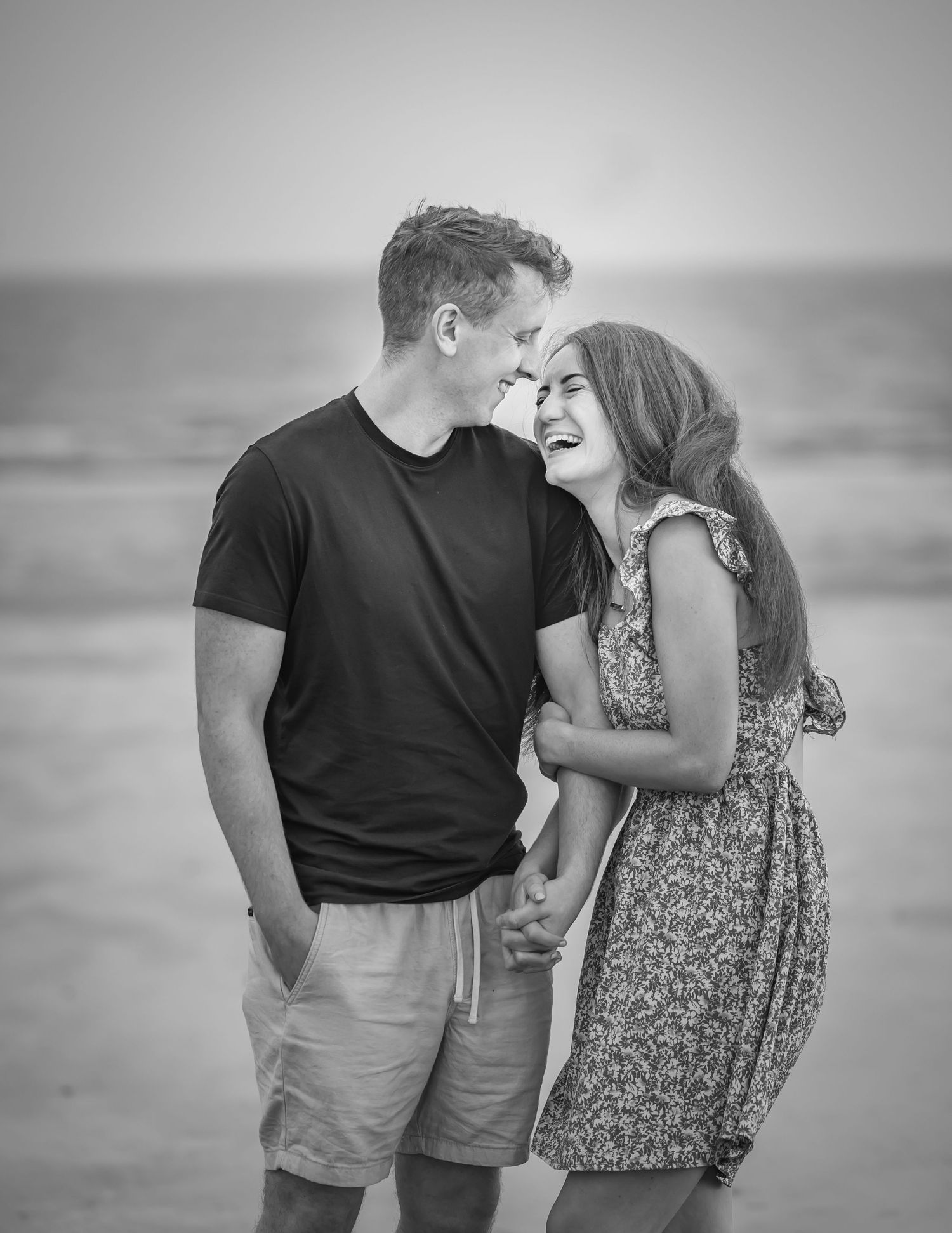 Black and white photo of a laughing couple holding hands and sharing a romantic moment on a beach.