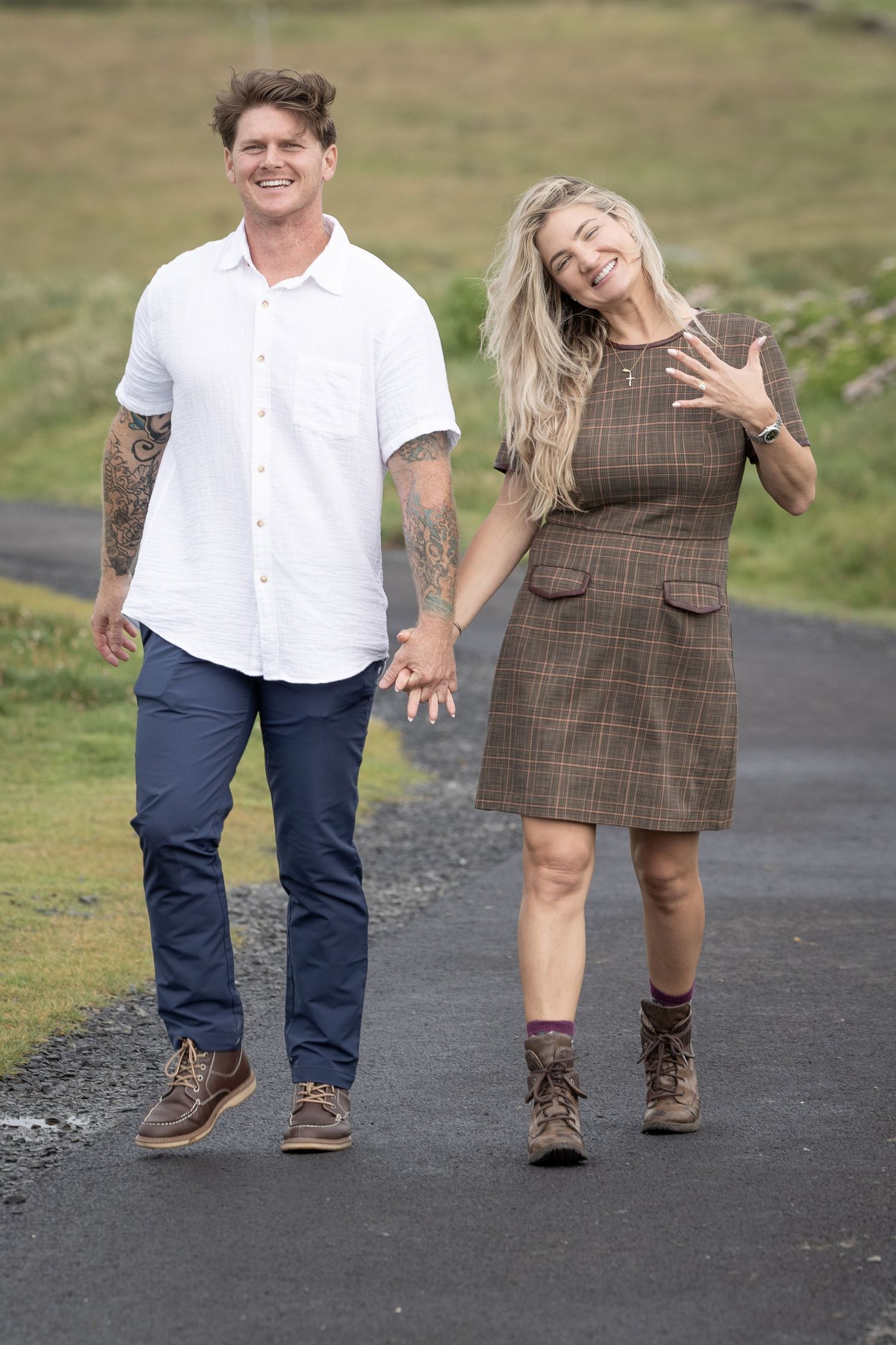 Engaged couple walks on rural path, woman showing ring and smiling, man in white shirt with tattoos beside her.