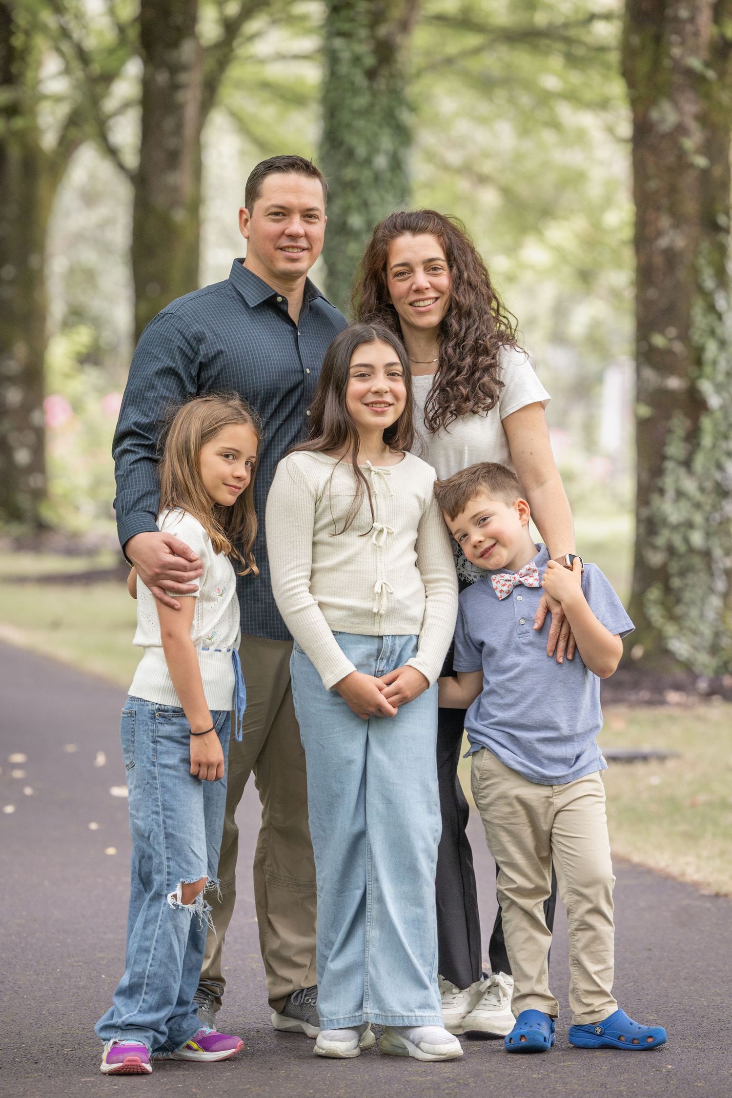 Happy family of five posing together on a tree-lined path, smiling outdoors in a park setting.