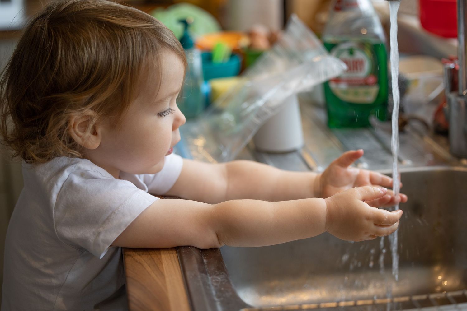 A small child reaches up to play with running water from a kitchen faucet.