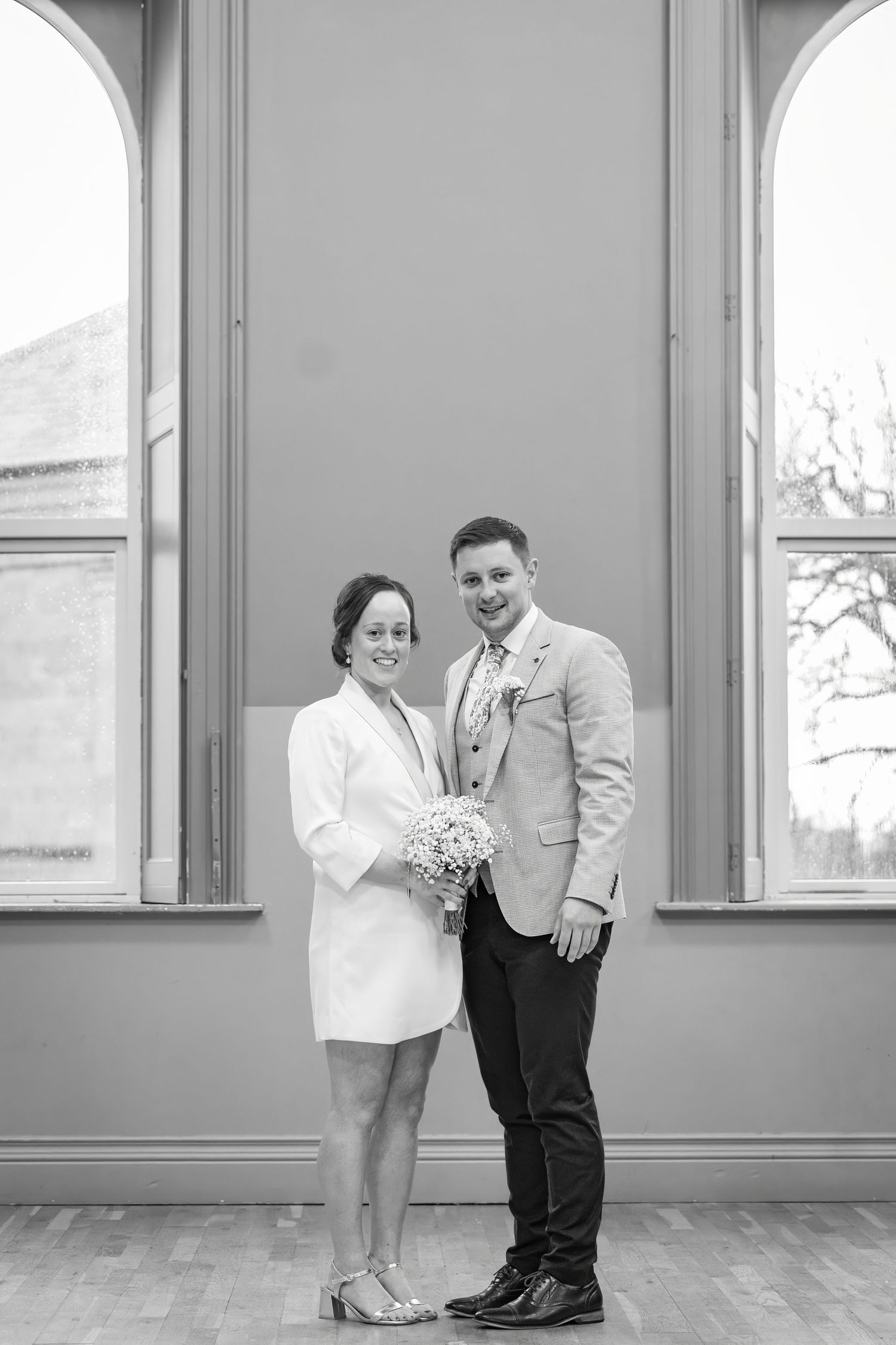 Black and white photo of a smiling couple posing indoors, bride holding bouquet in white dress, groom in light blazer.