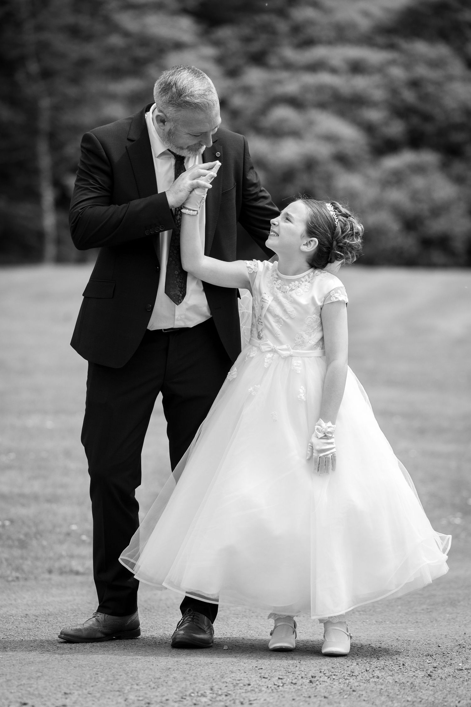 Black and white photo of a man in a suit kissing a flower girls hand at a wedding outdoors.