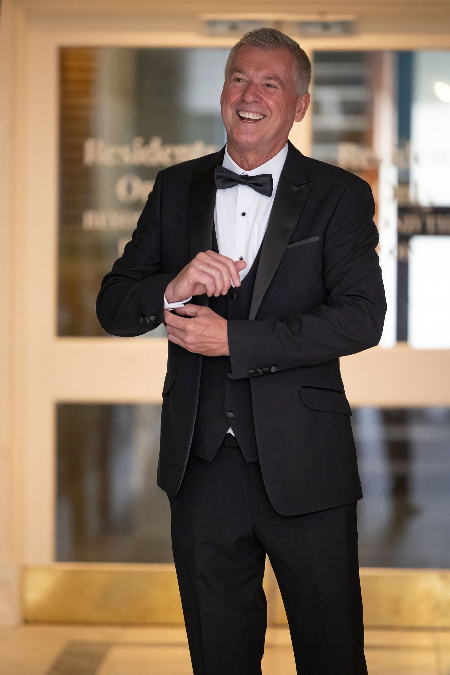 Smiling man in black tuxedo with bow tie adjusting cufflinks outside a hotel entrance.