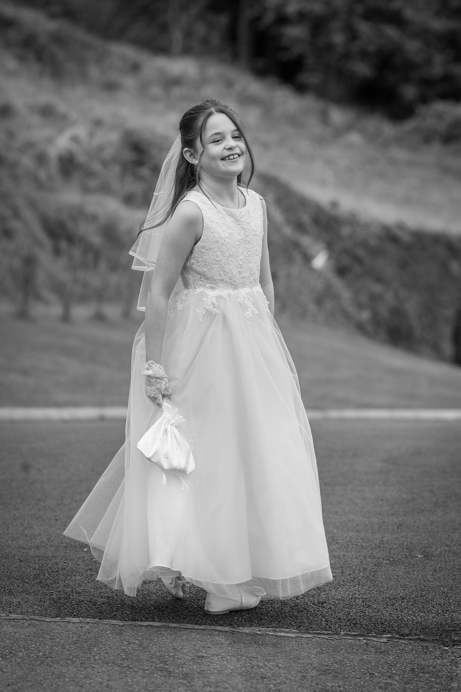 Young girl in white First Communion dress and veil smiles joyfully outdoors in black and white photo.