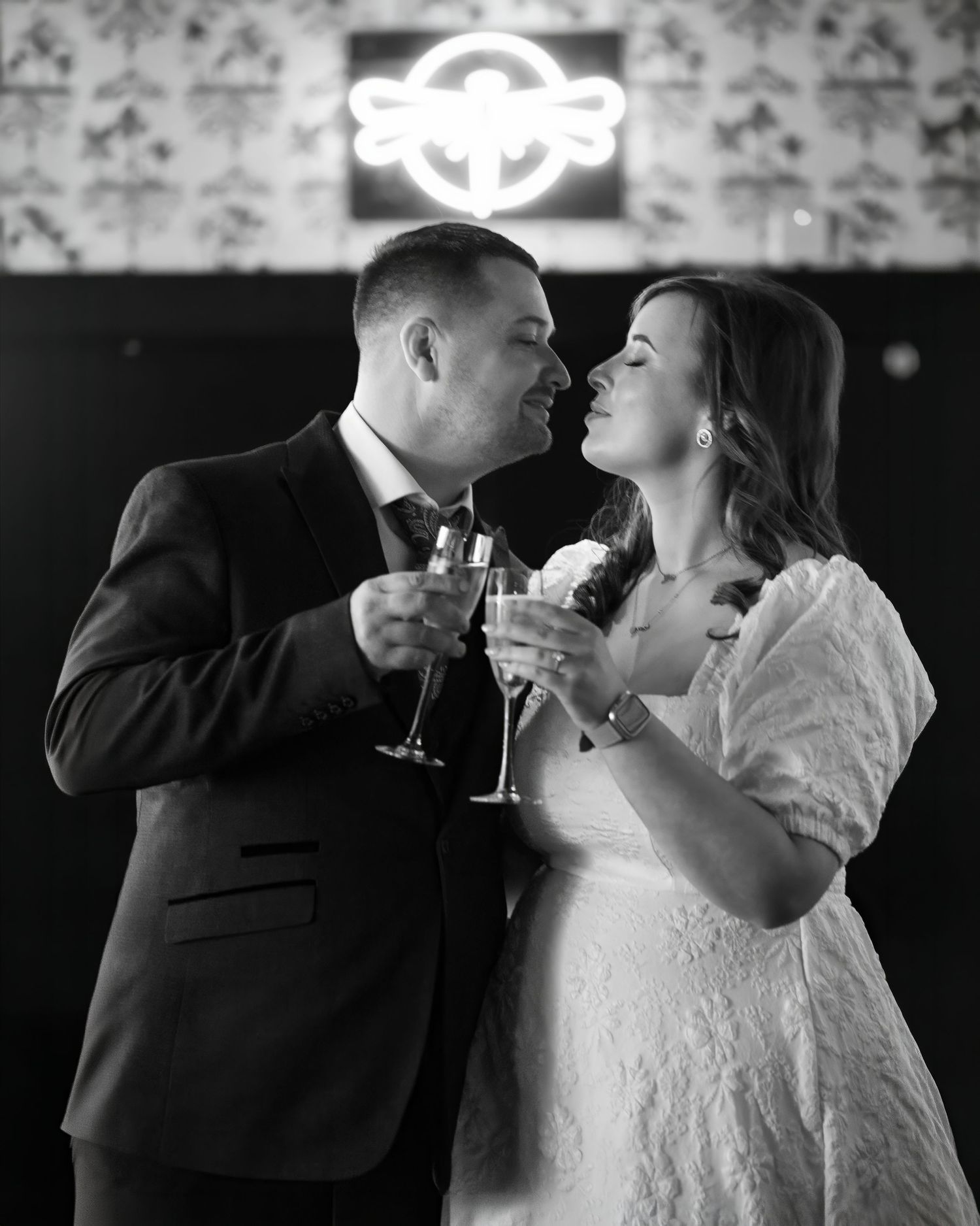 Black and white photo of a couple in wedding attire toasting with champagne glasses, smiling at each other romantically.