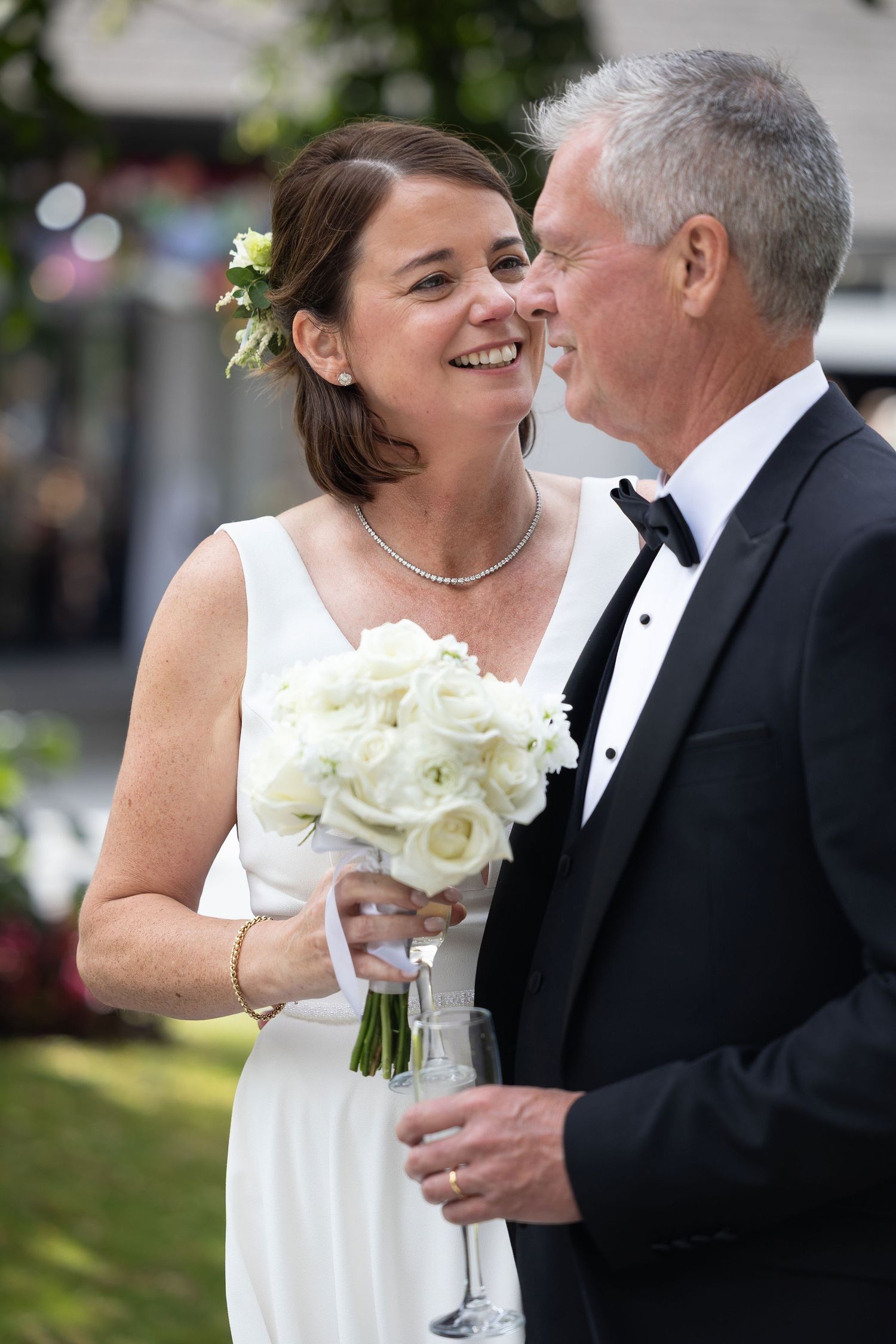 Smiling bride in white dress holding bouquet with groom in tuxedo at outdoor wedding ceremony.