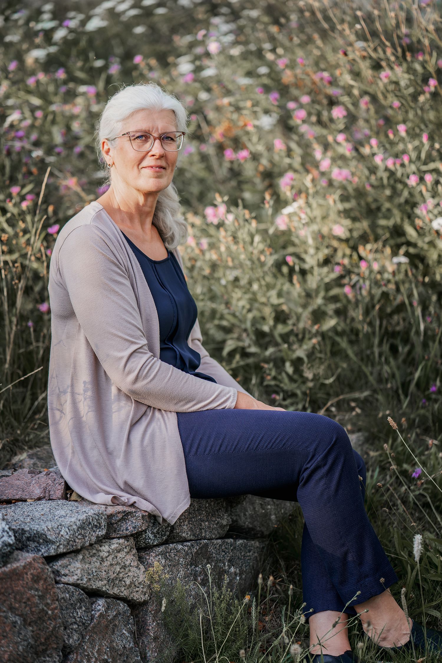 Person seated on stone wall surrounded by pink wildflowers in garden setting.