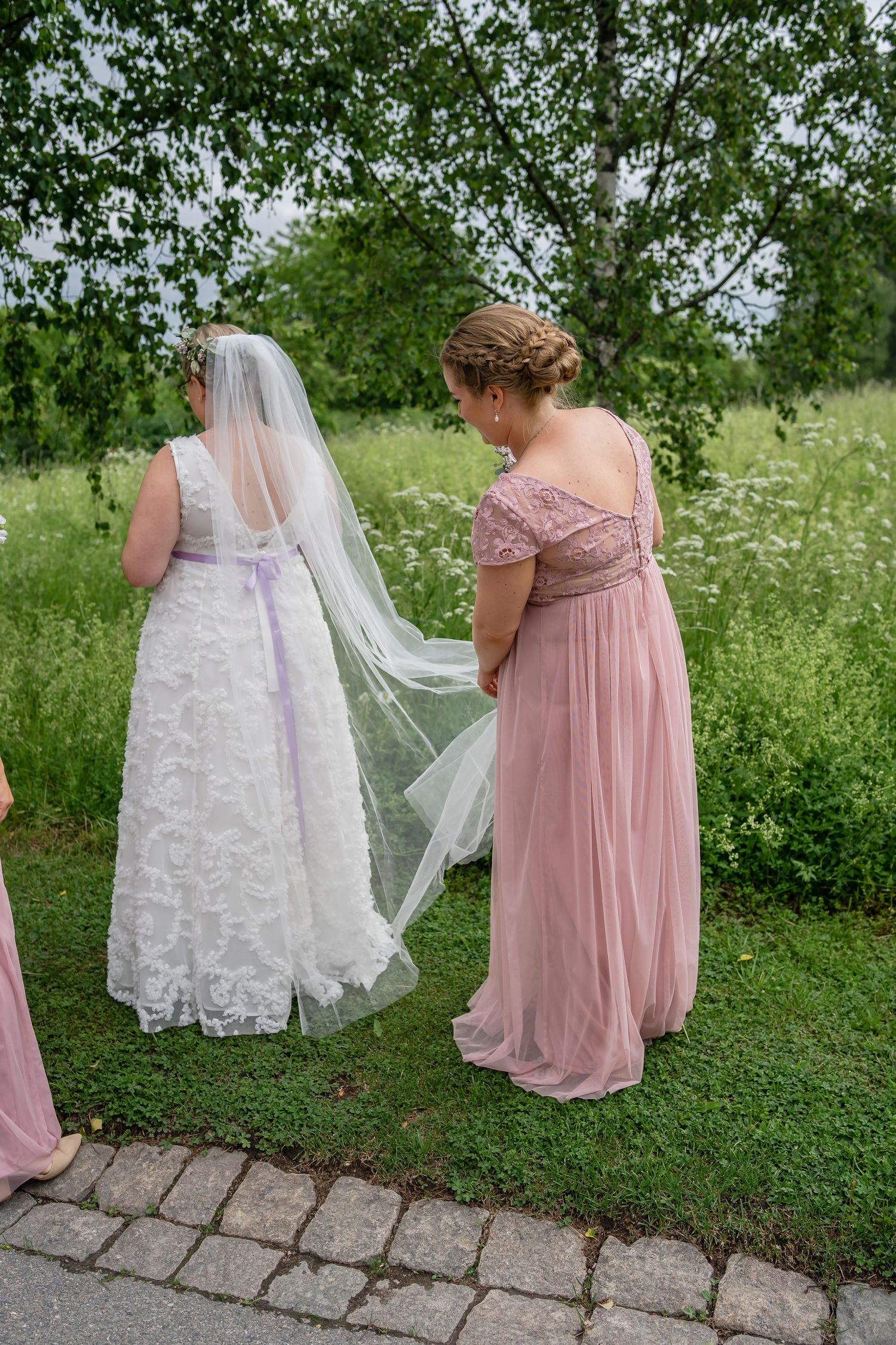 Bride in white lace dress with flowing veil assisted by bridesmaid in pink gown.