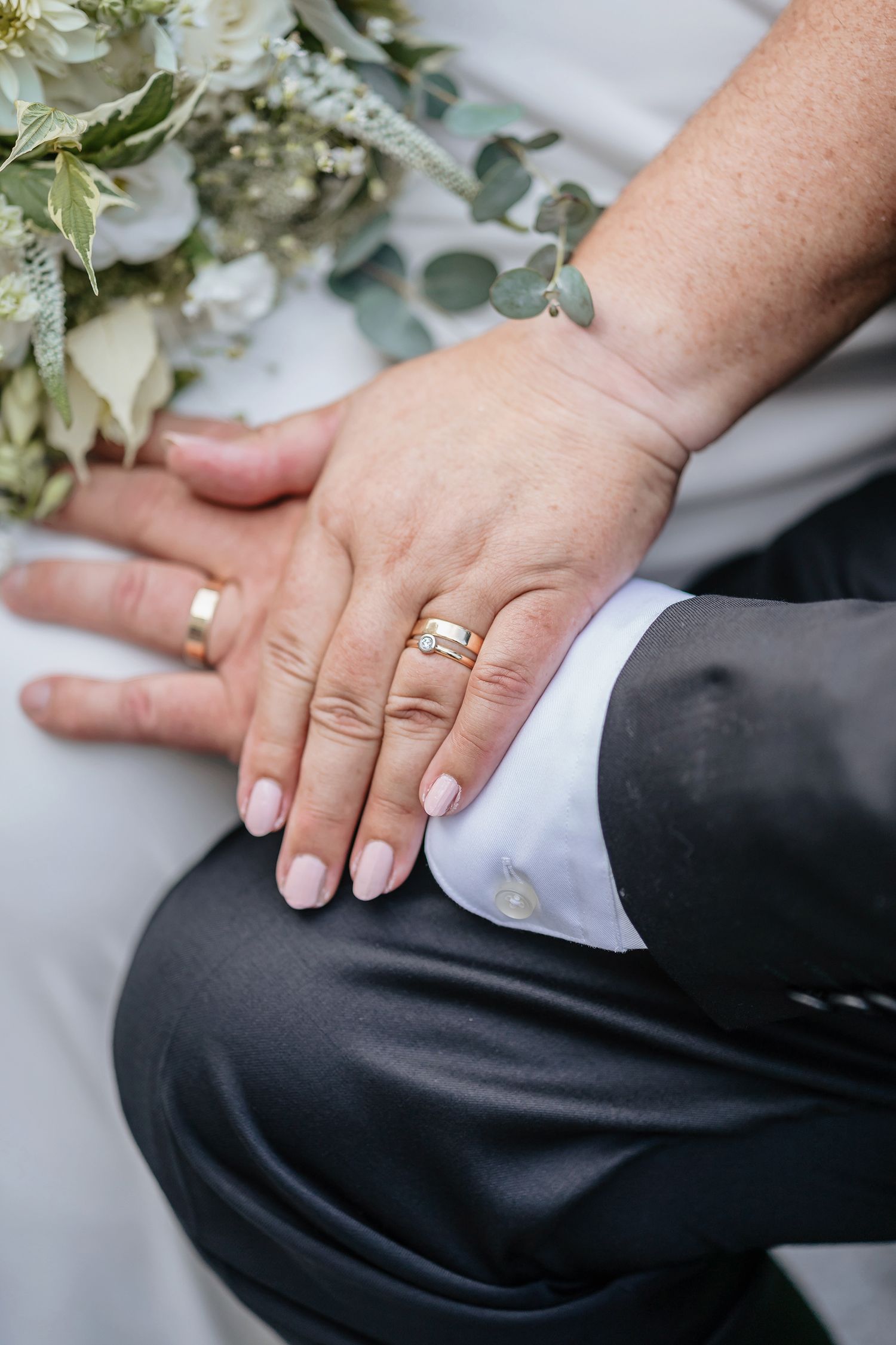 Close-up of newlywed couple's hands with wedding rings and white floral bouquet against formal attire.