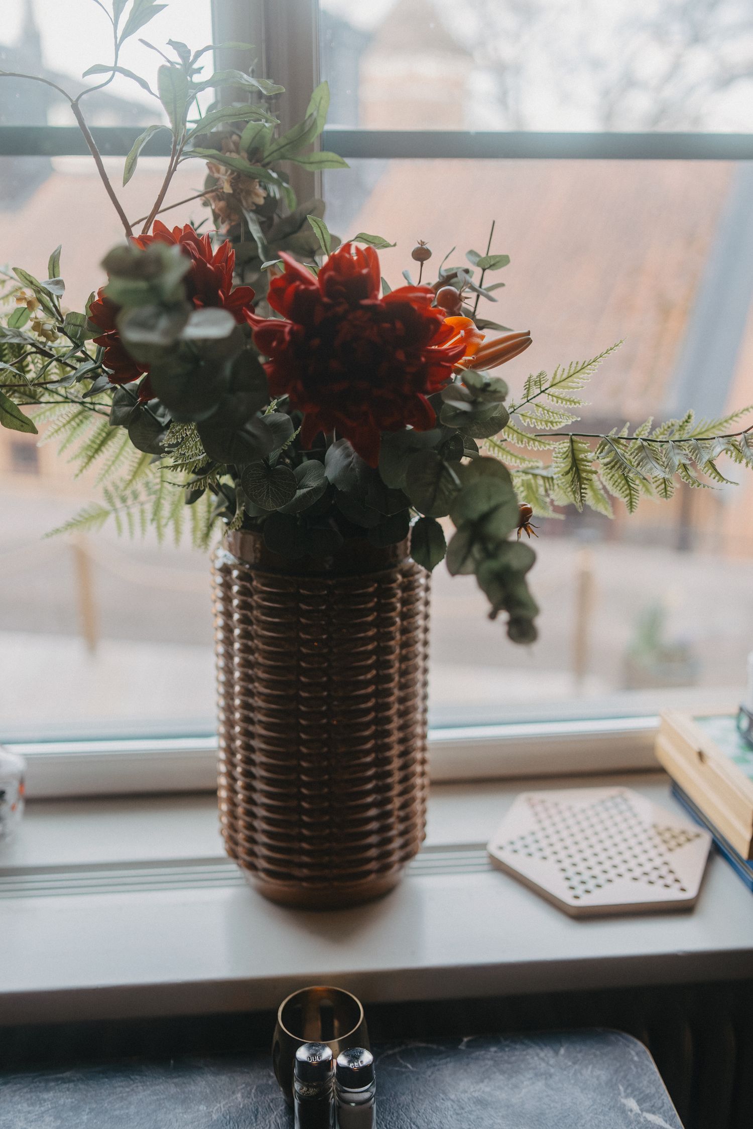 Red floral arrangement with ferns in brown textured vase by window.