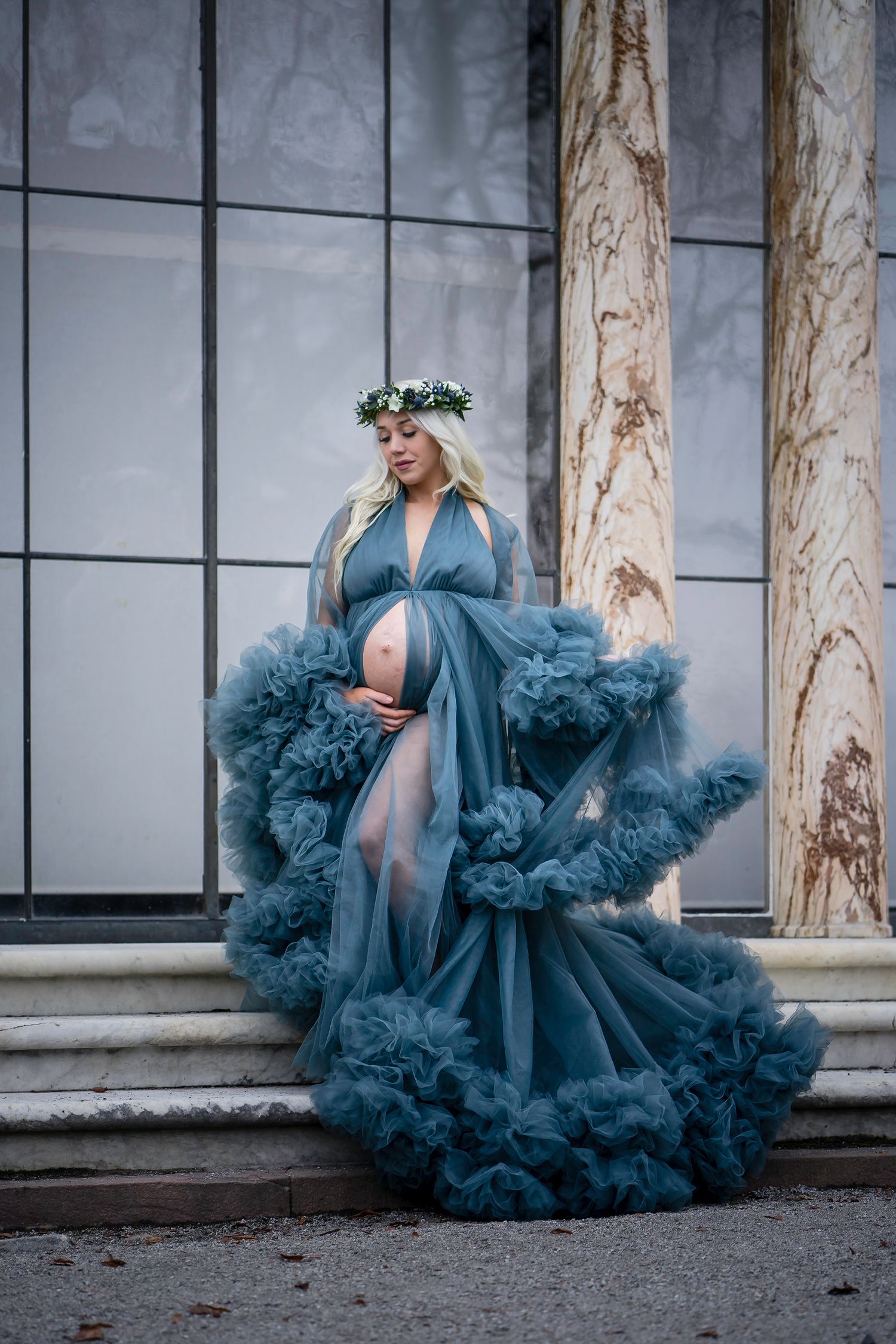 A pregnant woman poses in a flowing blue tulle gown and floral crown on marble steps against a modern building exterior.
