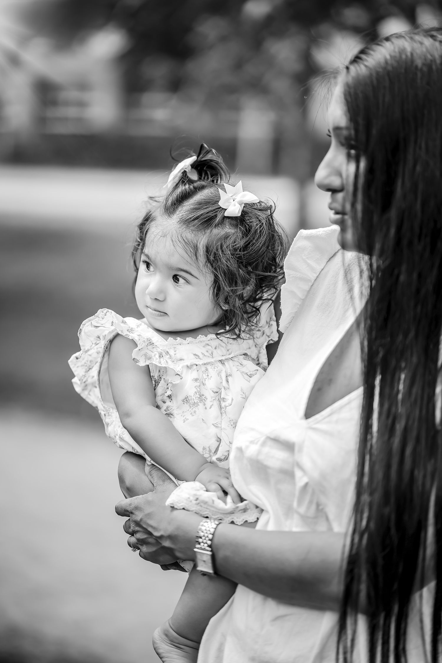 Black and white portrait of a toddler in a ruffled dress being held by someone with long dark hair.