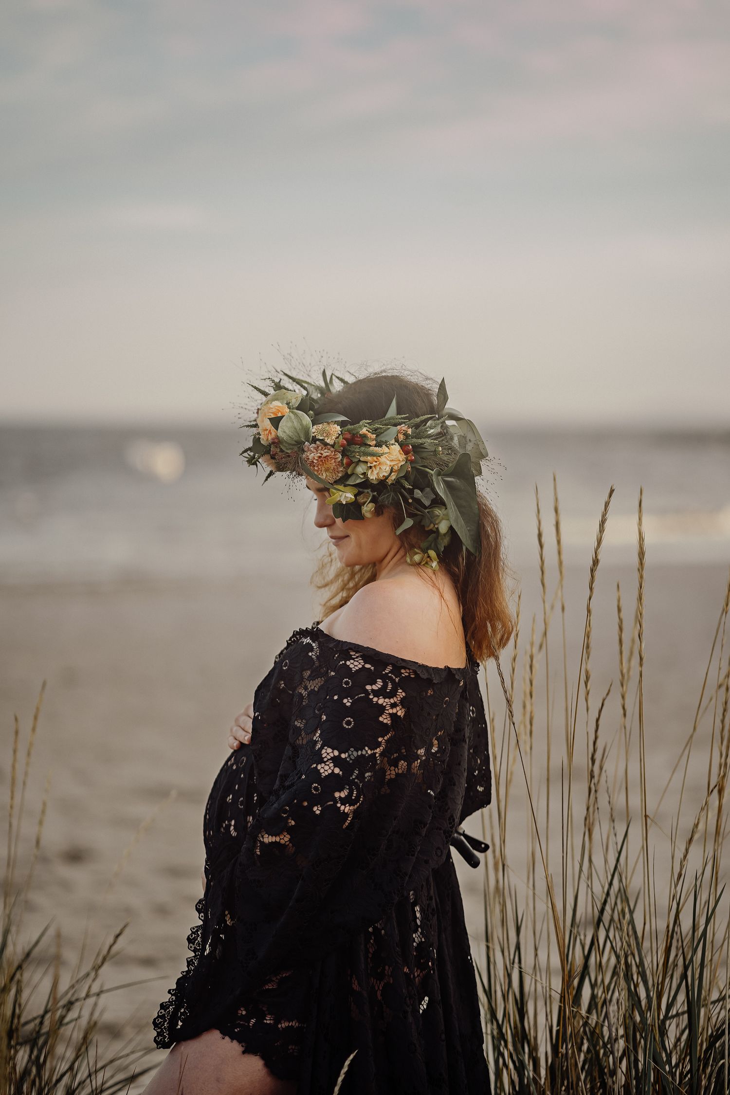 Woman in black lace dress wearing floral crown stands on beach at sunset with sea grass in foreground.