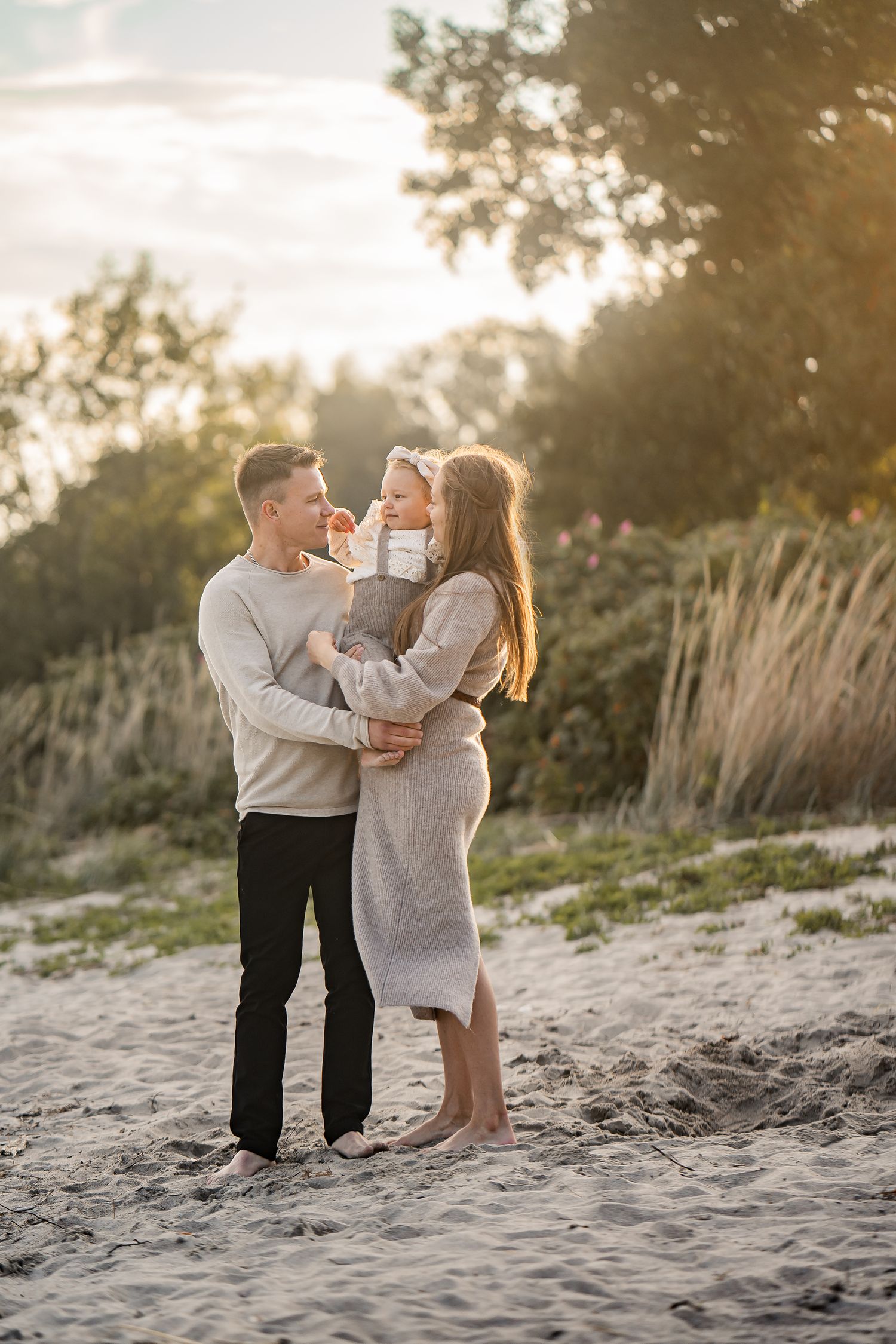 A couple and small child share a tender moment together on a sandy beach at sunset with tall grass in the background.