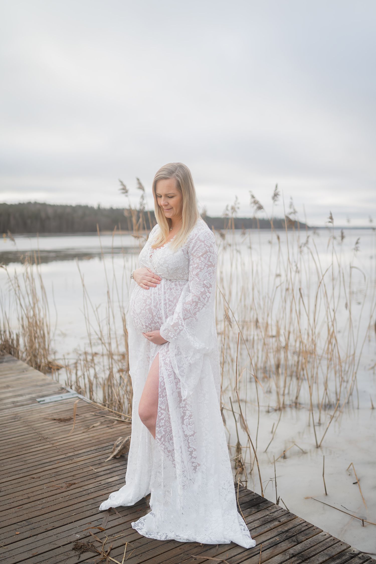 Woman in white lace dress standing on wooden dock by lake during winter.