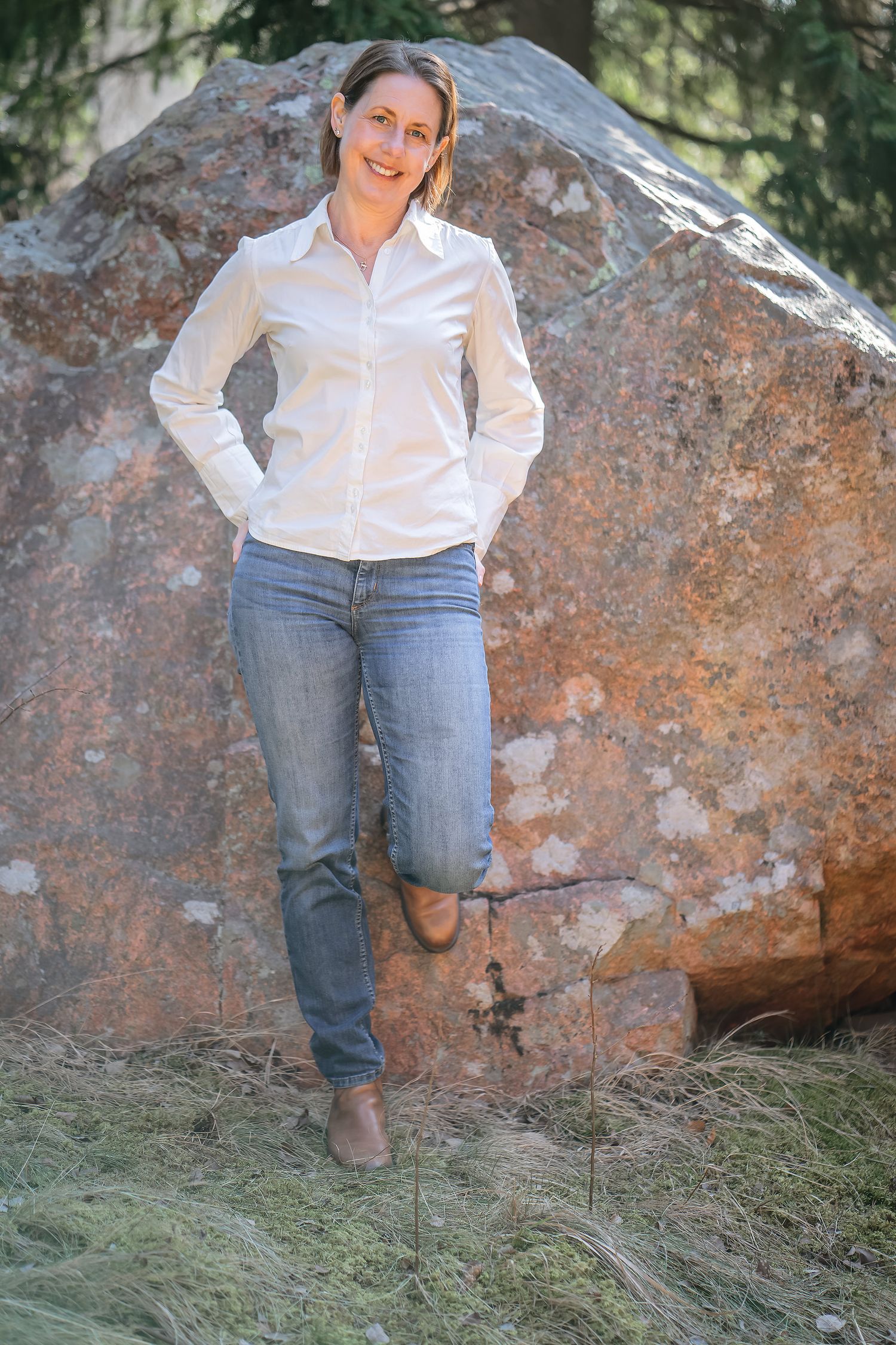Person in white button-down shirt and jeans leaning against large boulder outdoors.