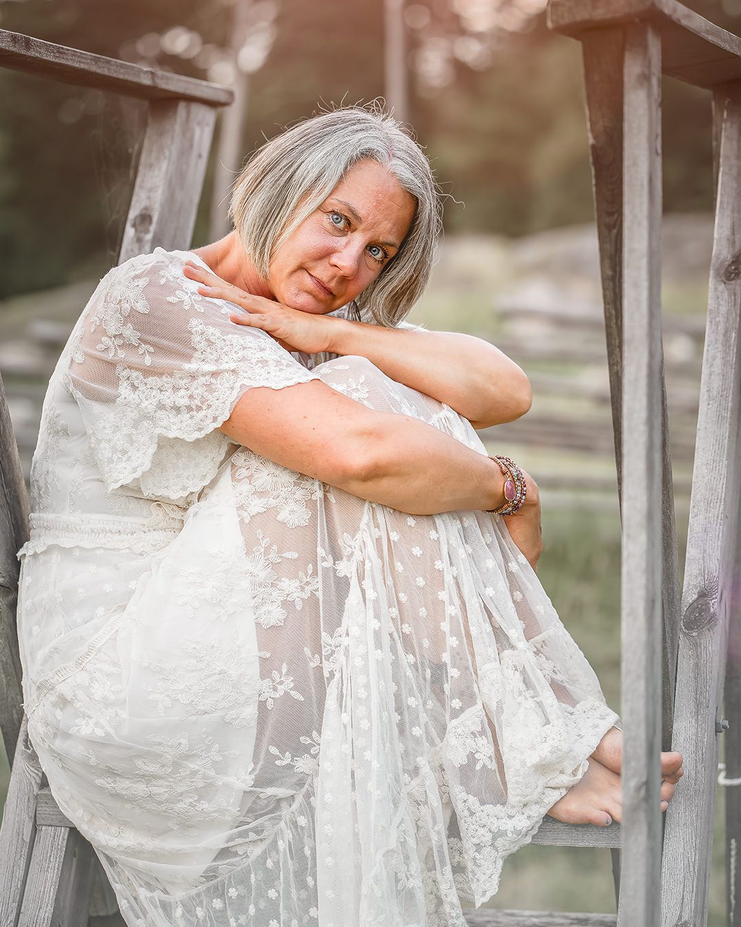 Dreamy portrait in a white lace dress shows someone sitting on wooden steps in soft natural lighting.