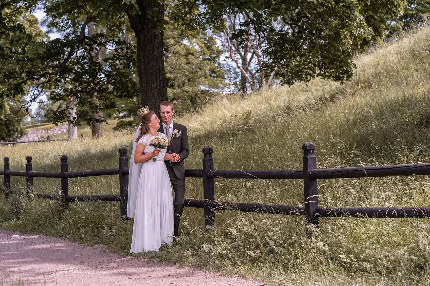 Couple stands together by rustic wood fence on hillside path during outdoor wedding photoshoot.