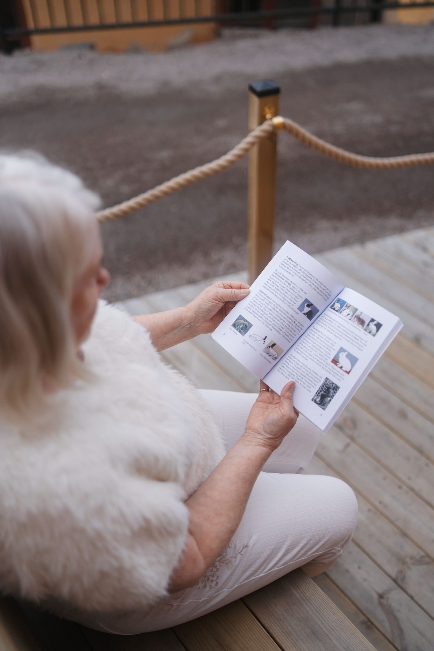 Someone in a white fluffy sweater reads a small booklet while sitting on wooden decking near a rope railing.