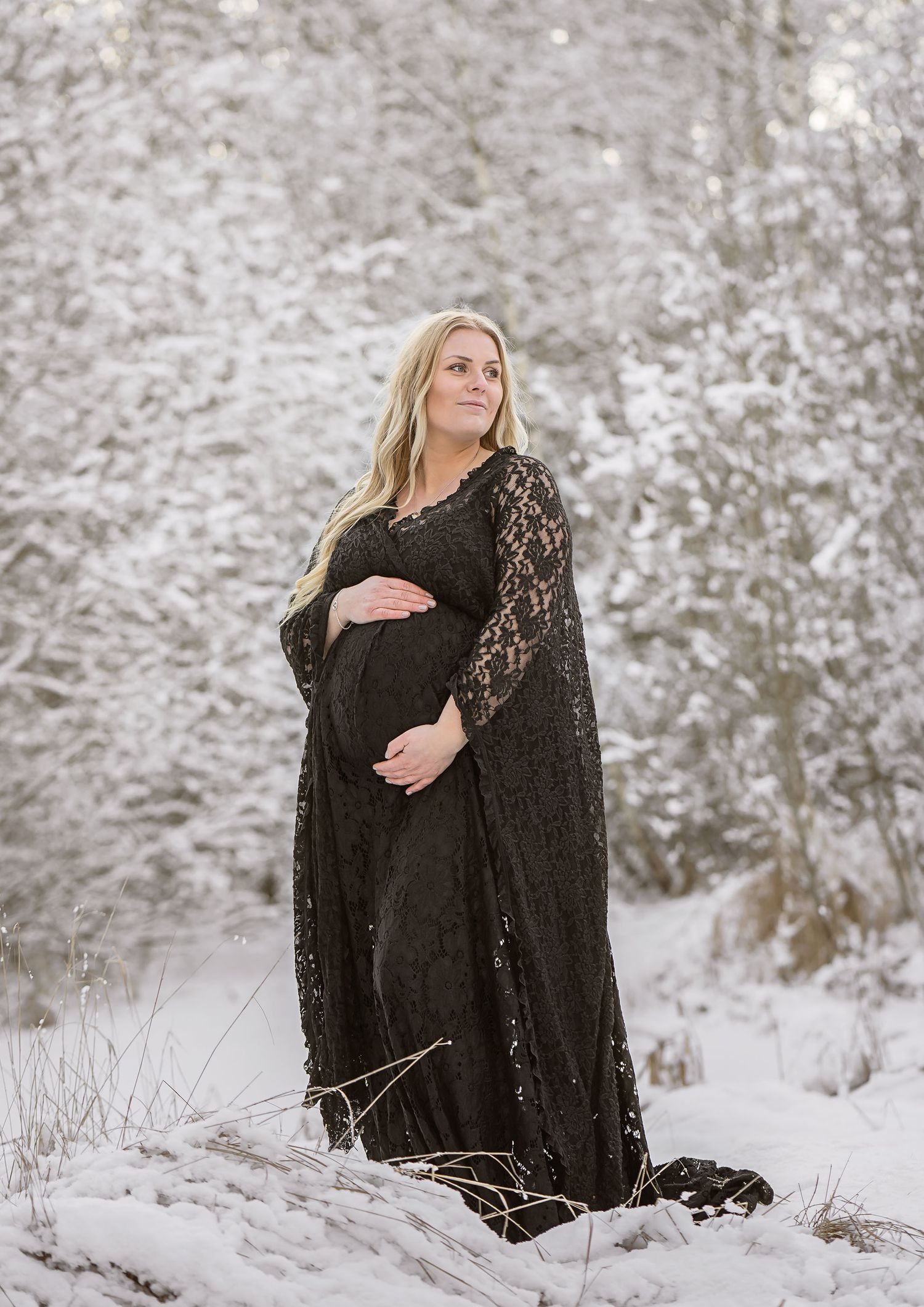 Maternity portrait in black lace dress against snowy winter landscape backdrop.