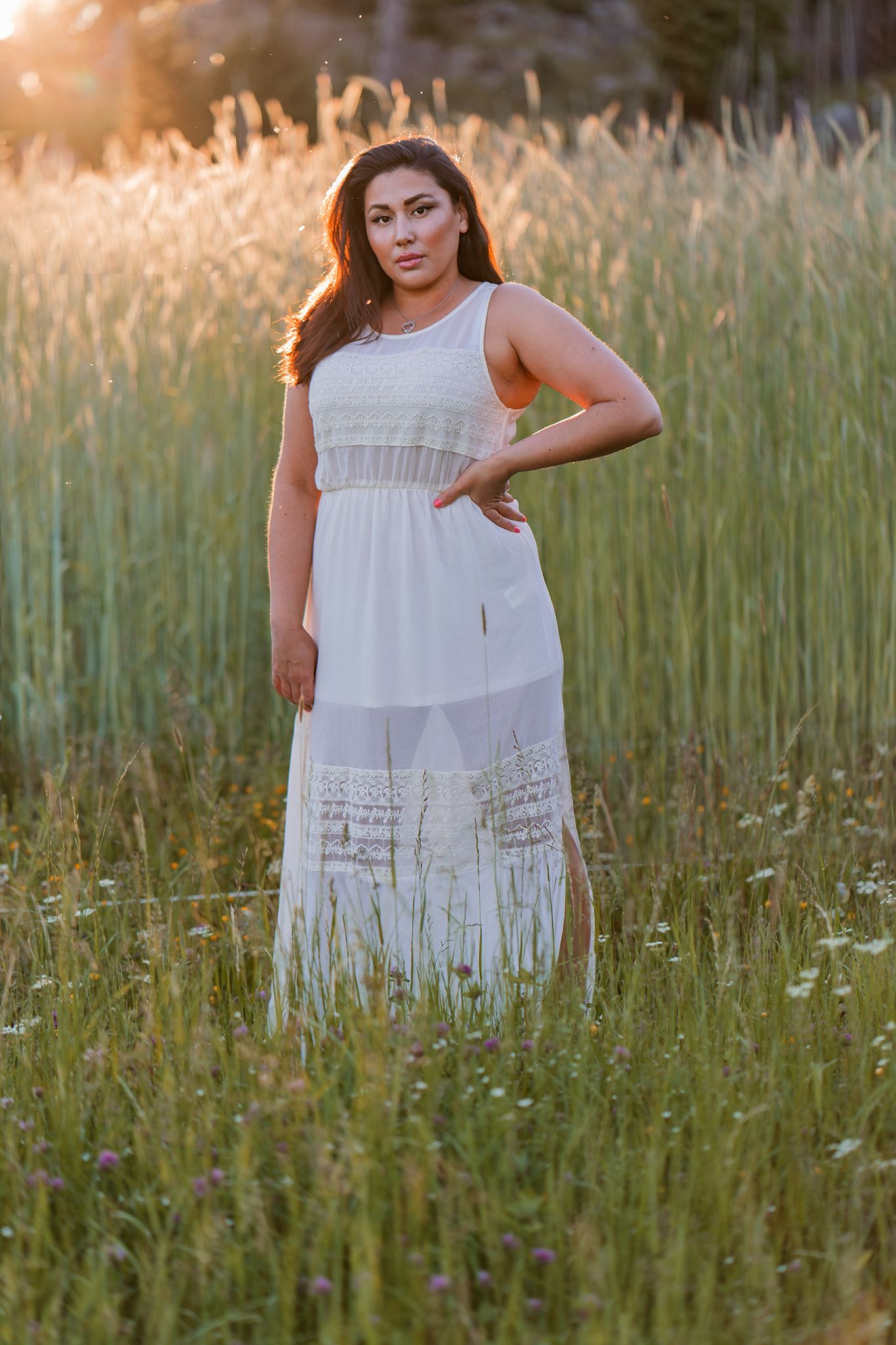 A person in a flowing white dress poses in a field of tall grass during golden hour sunset lighting.
