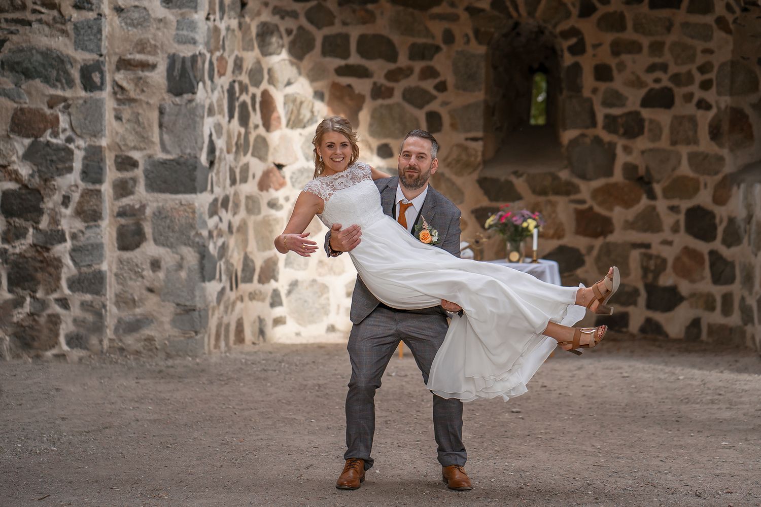 Joyful newlyweds share a playful moment against a stone wall during their wedding photoshoot.