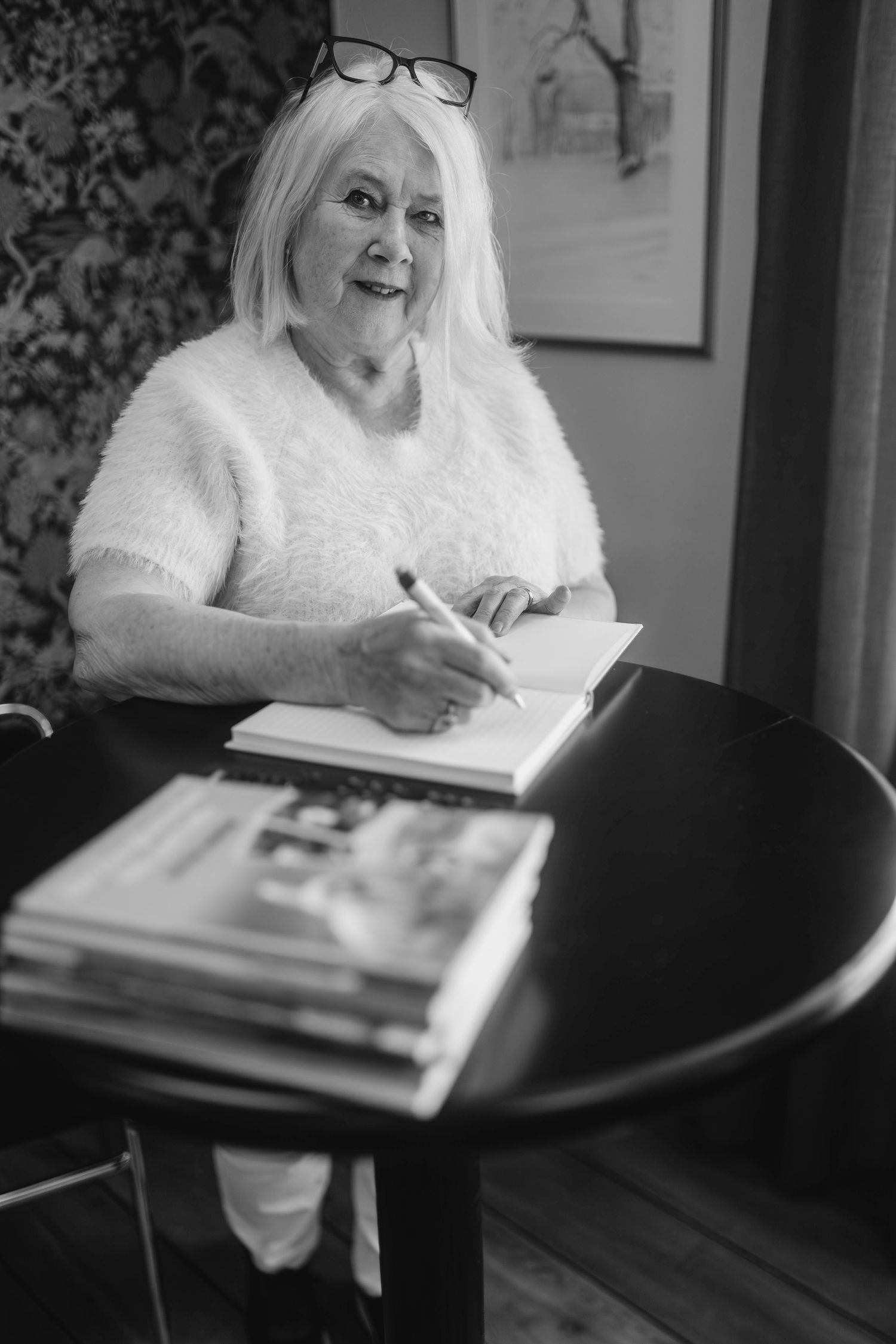 Someone writes in a notebook at a round table with reading glasses perched on their head.