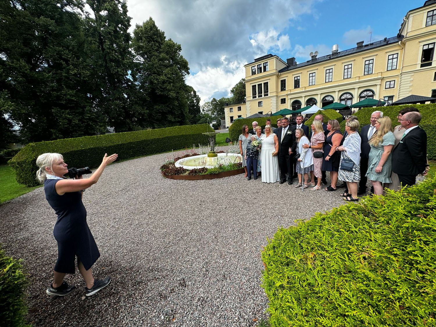 Photographer captures wedding party group photo outside elegant yellow mansion.