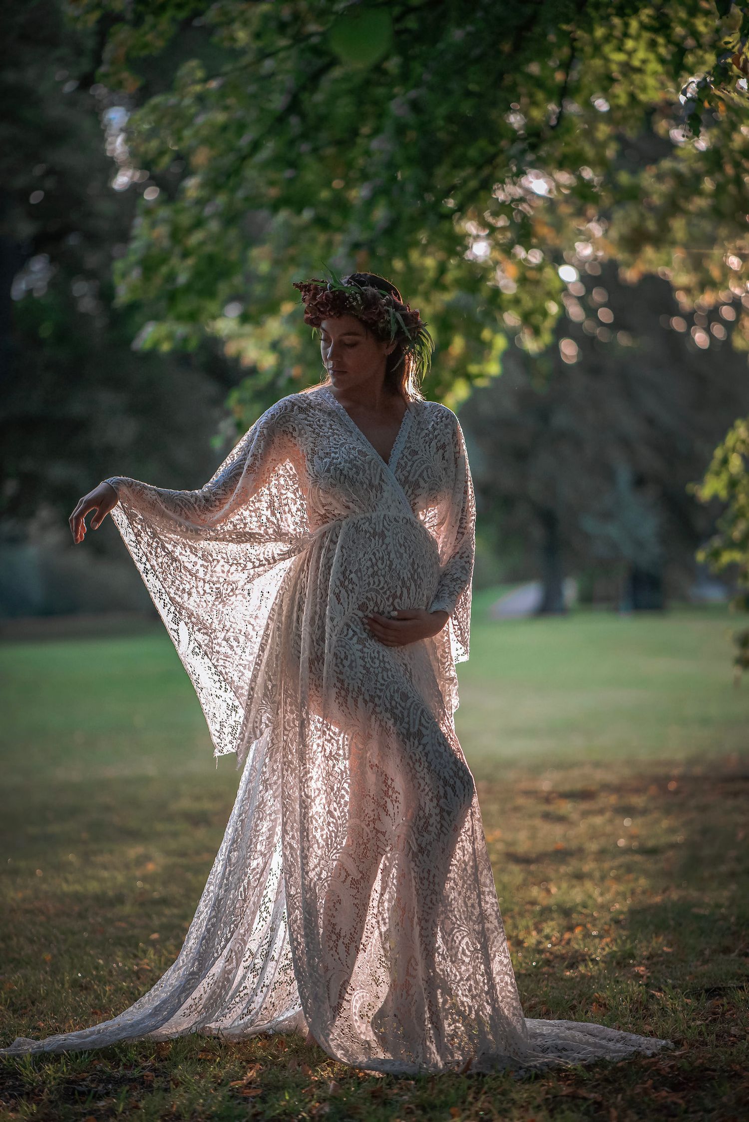 A person in a flowing lace maternity gown poses elegantly in a sunlit outdoor setting with trees in background.