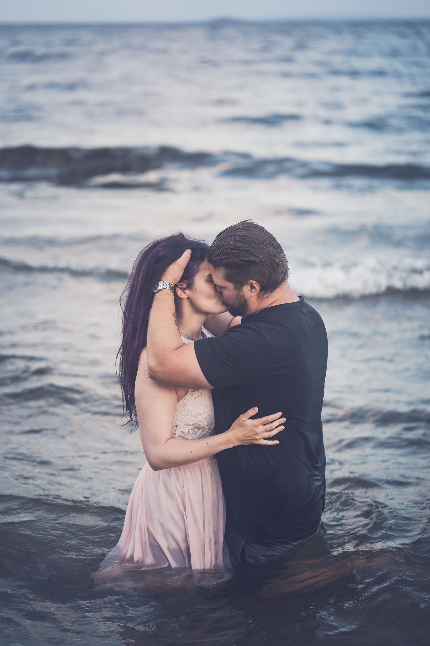 A romantic couple embraces and kisses while standing in ocean waves at dusk.