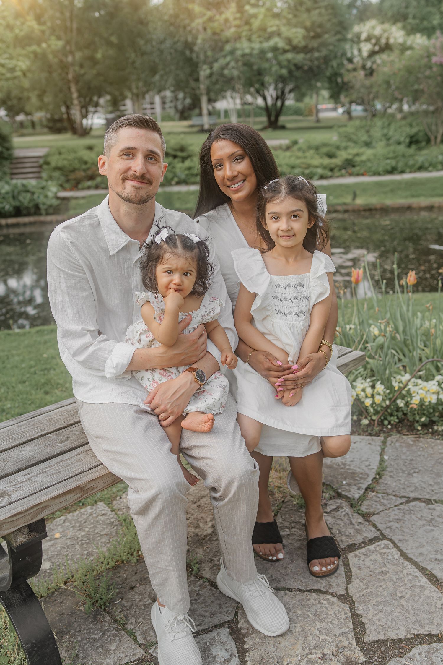 Family wearing coordinated white outfits poses together on a park bench during a sunny outdoor photo session.