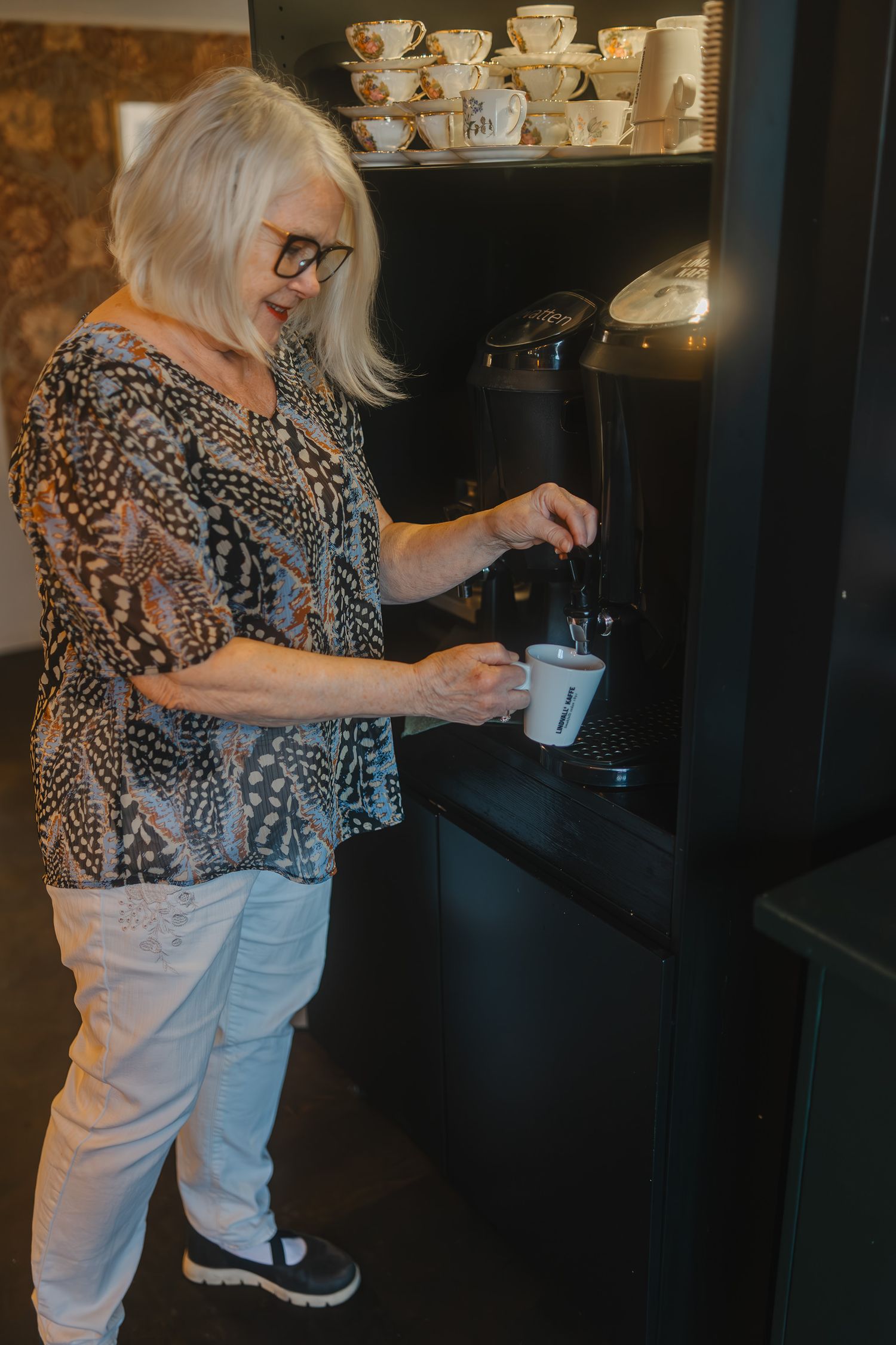 Person making coffee at home with collection of teacups displayed on shelves.