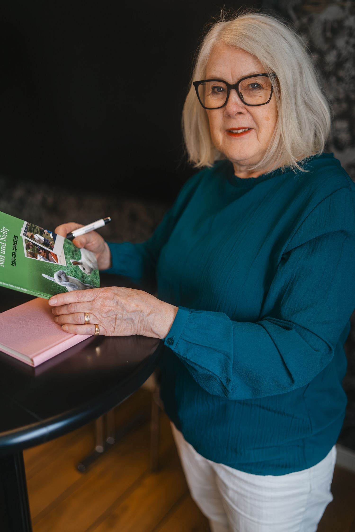Senior woman in teal sweater opening a green book at home.