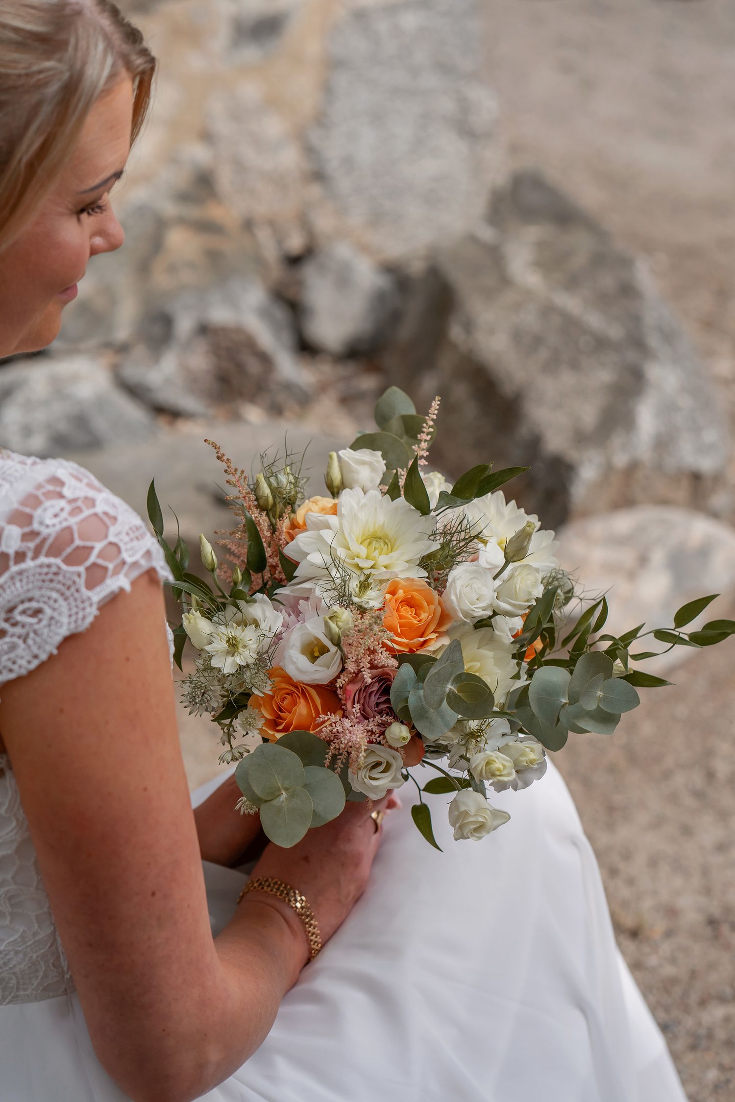 A bride holds a beautiful bouquet featuring white roses, orange flowers, and eucalyptus against stone wall.