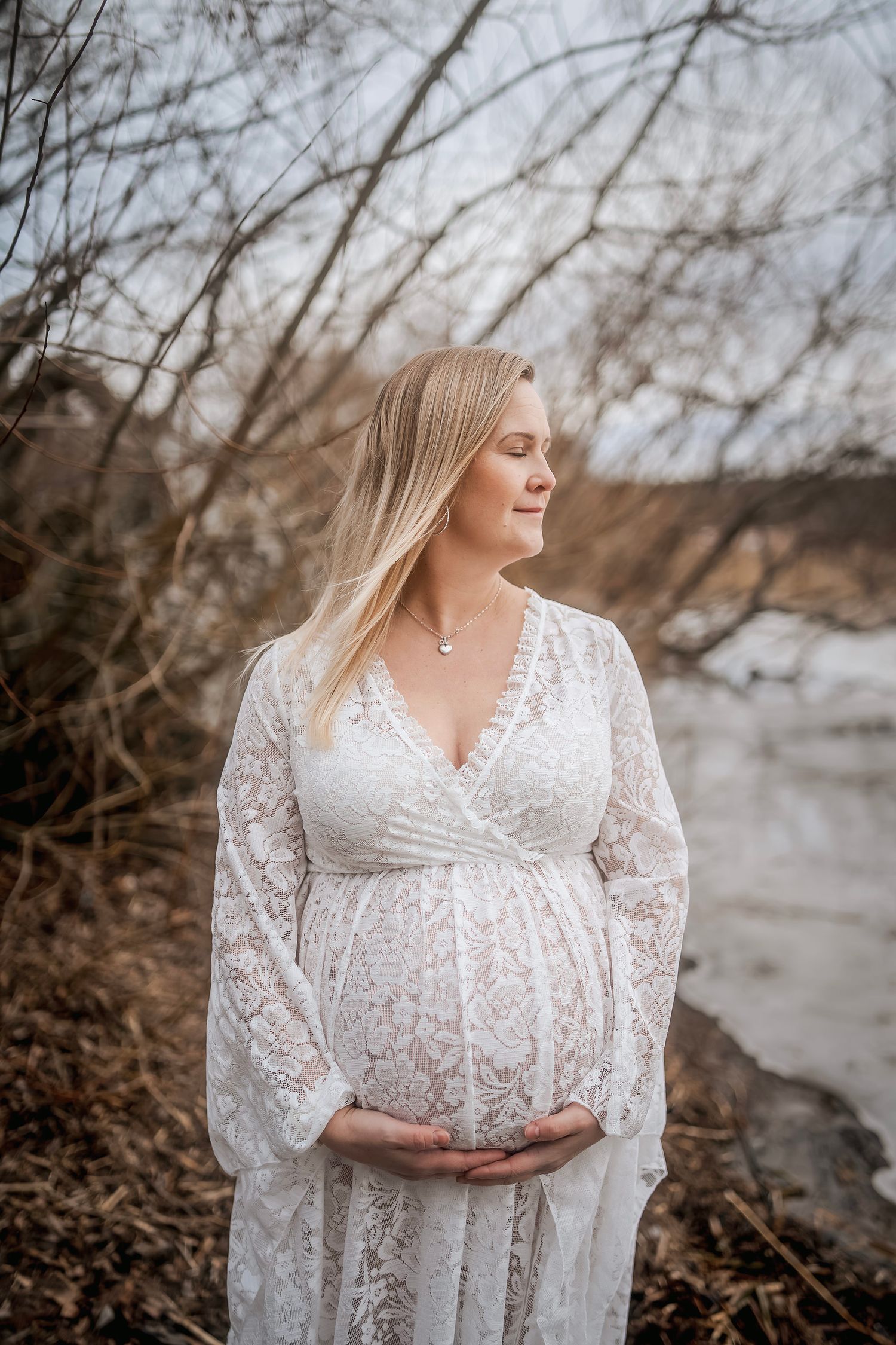 A person in a white lace maternity dress poses outdoors near a lake.
