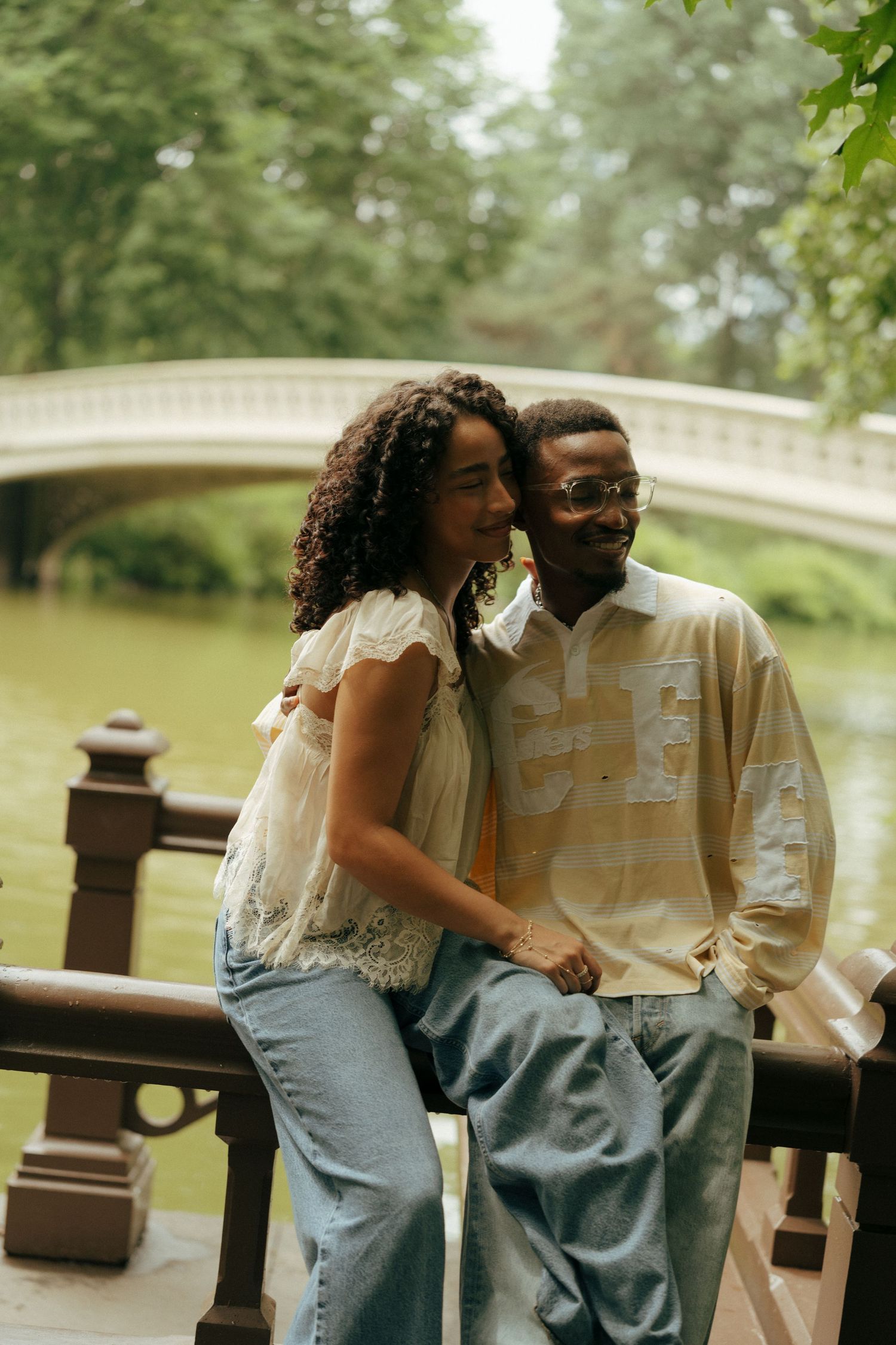 A couple poses romantically near Bow Bridge in Central Park, surrounded by lush greenery and a serene lake.